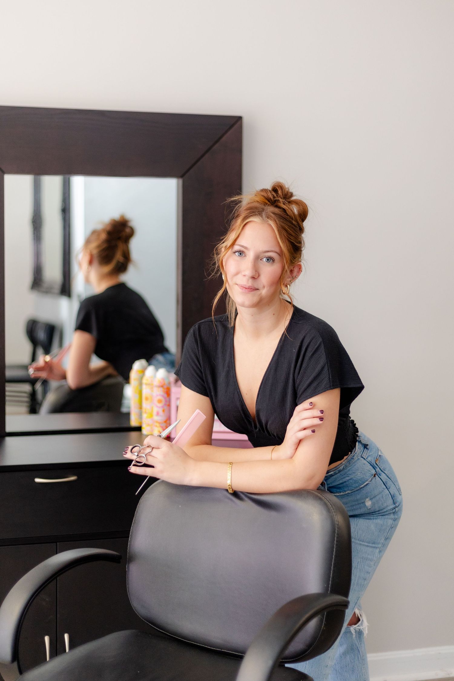 Red-haired salon professional leaning on styling chair in front of mirror holding hair products.