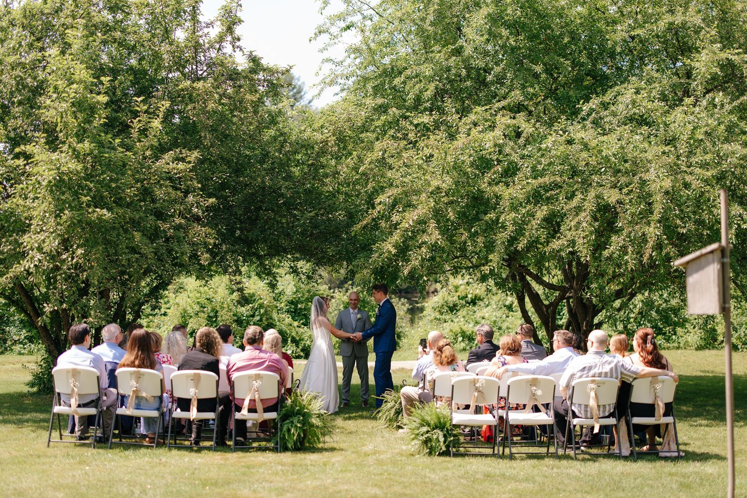 Intimate outdoor wedding ceremony with couple at altar surrounded by seated guests under green trees.