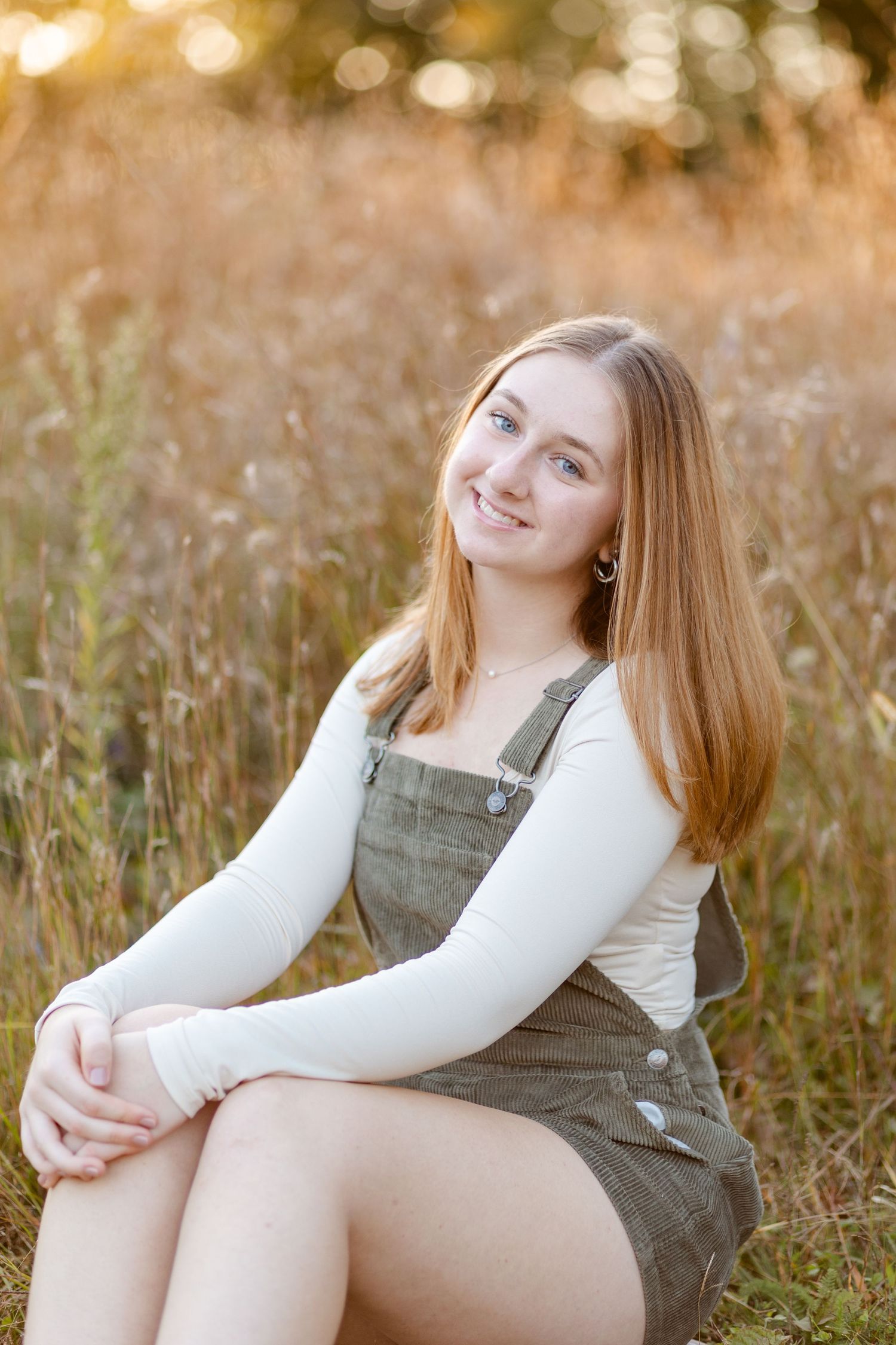 Smiling teenage girl in olive pinafore dress sitting in golden sunlit field during sunset.