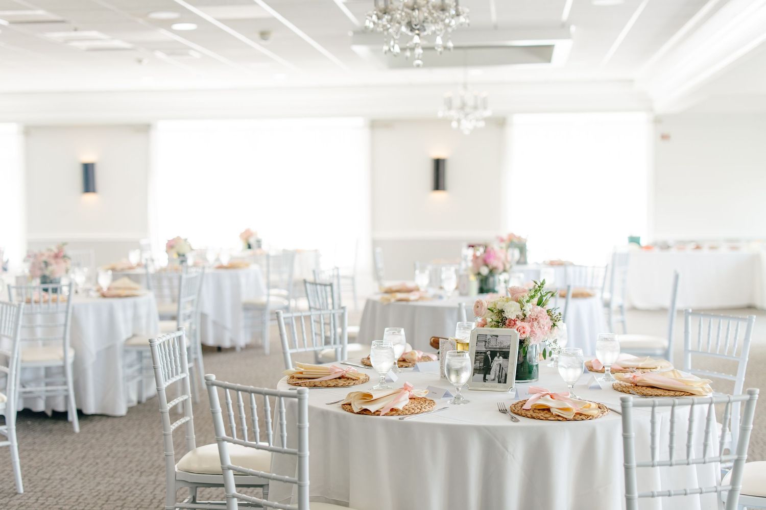 Elegant wedding reception ballroom with white round tables, chiavari chairs, and pink floral centerpieces.