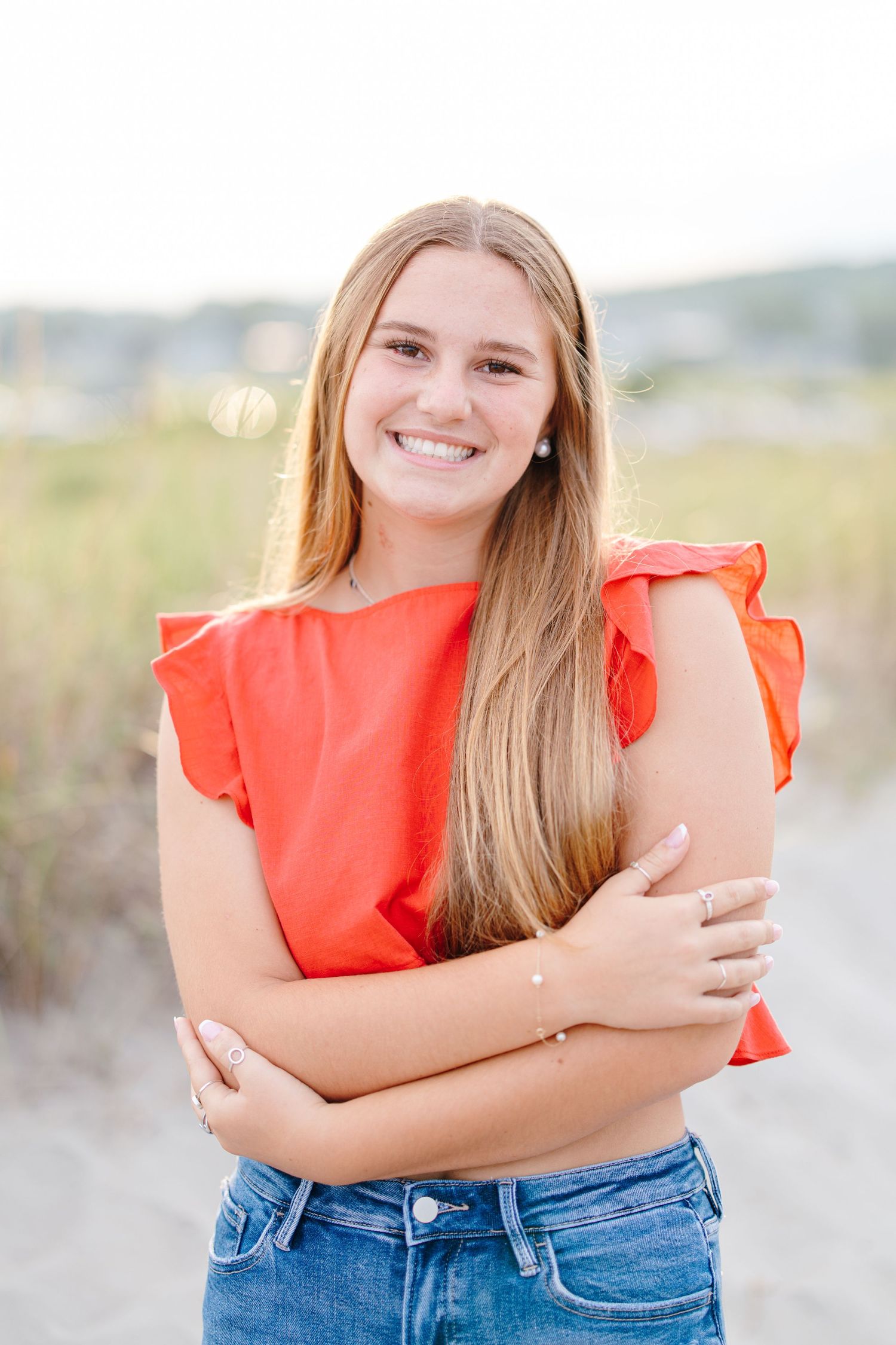Smiling teen girl in orange ruffled top with jeans poses on sandy beach at sunset.