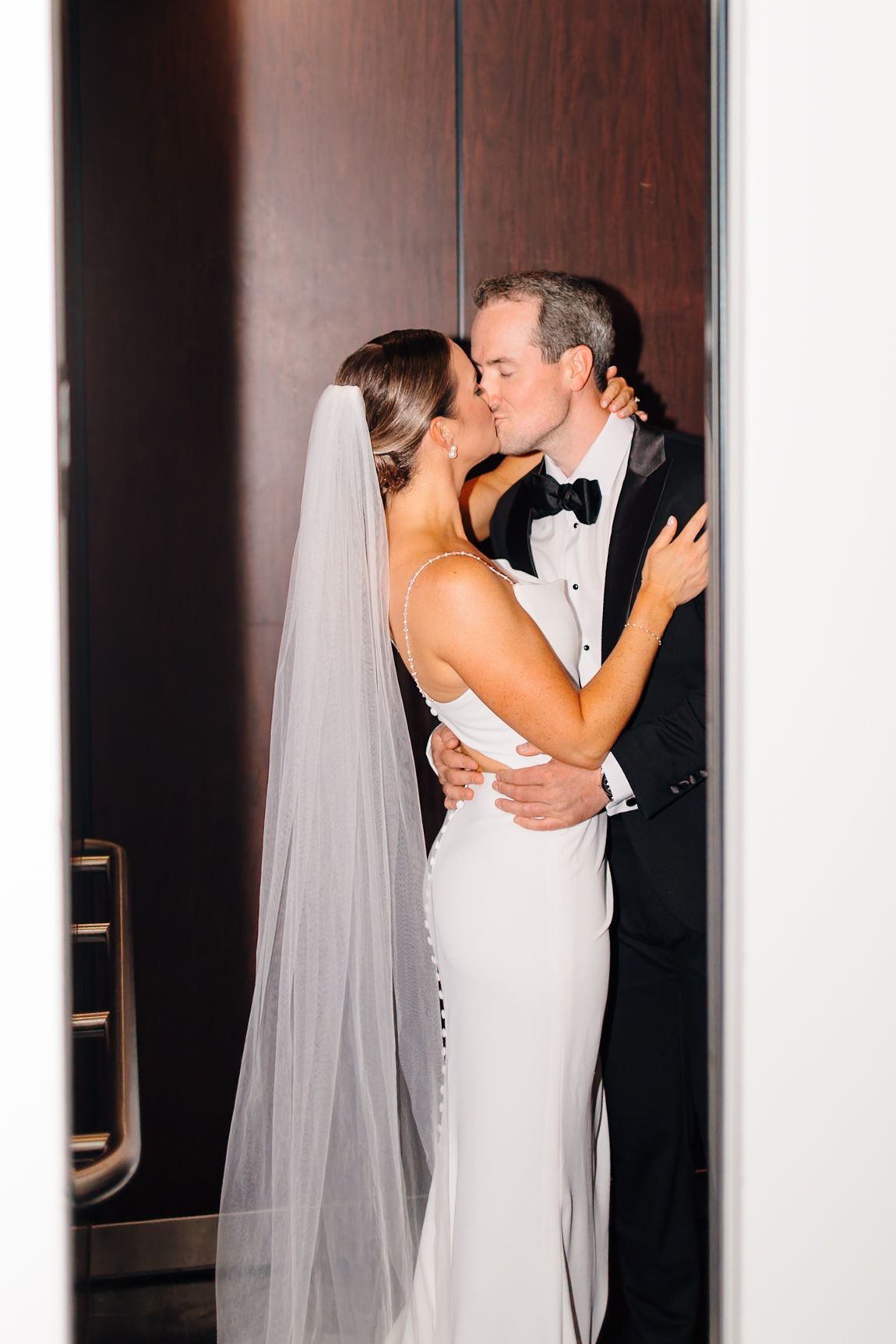 Bride and groom share a romantic kiss in a doorway, she wearing a white gown with veil, he in a black tuxedo.