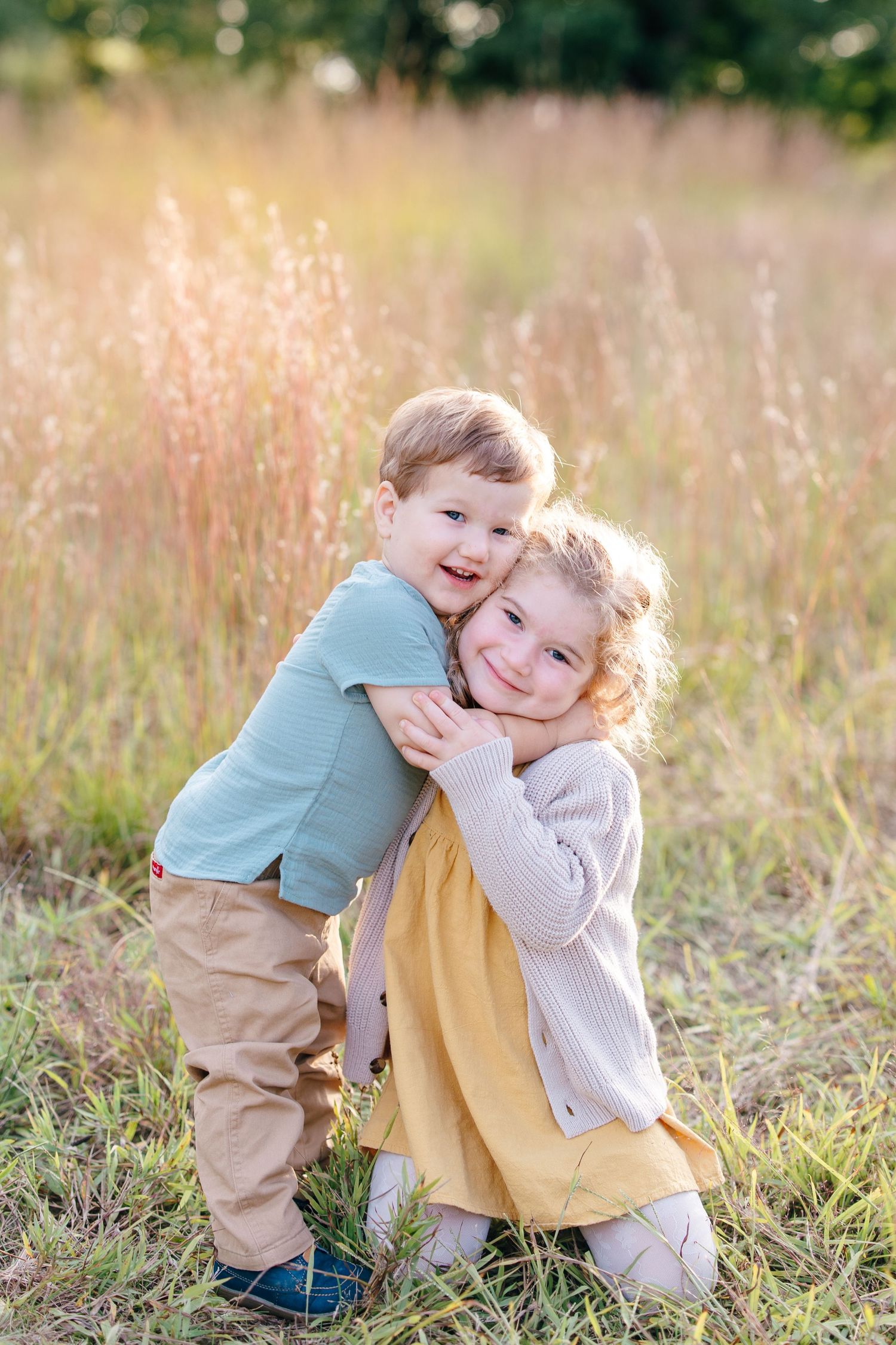 Two young siblings share a sweet embrace in a sunlit field, with the older boy hugging his little sister from behind.