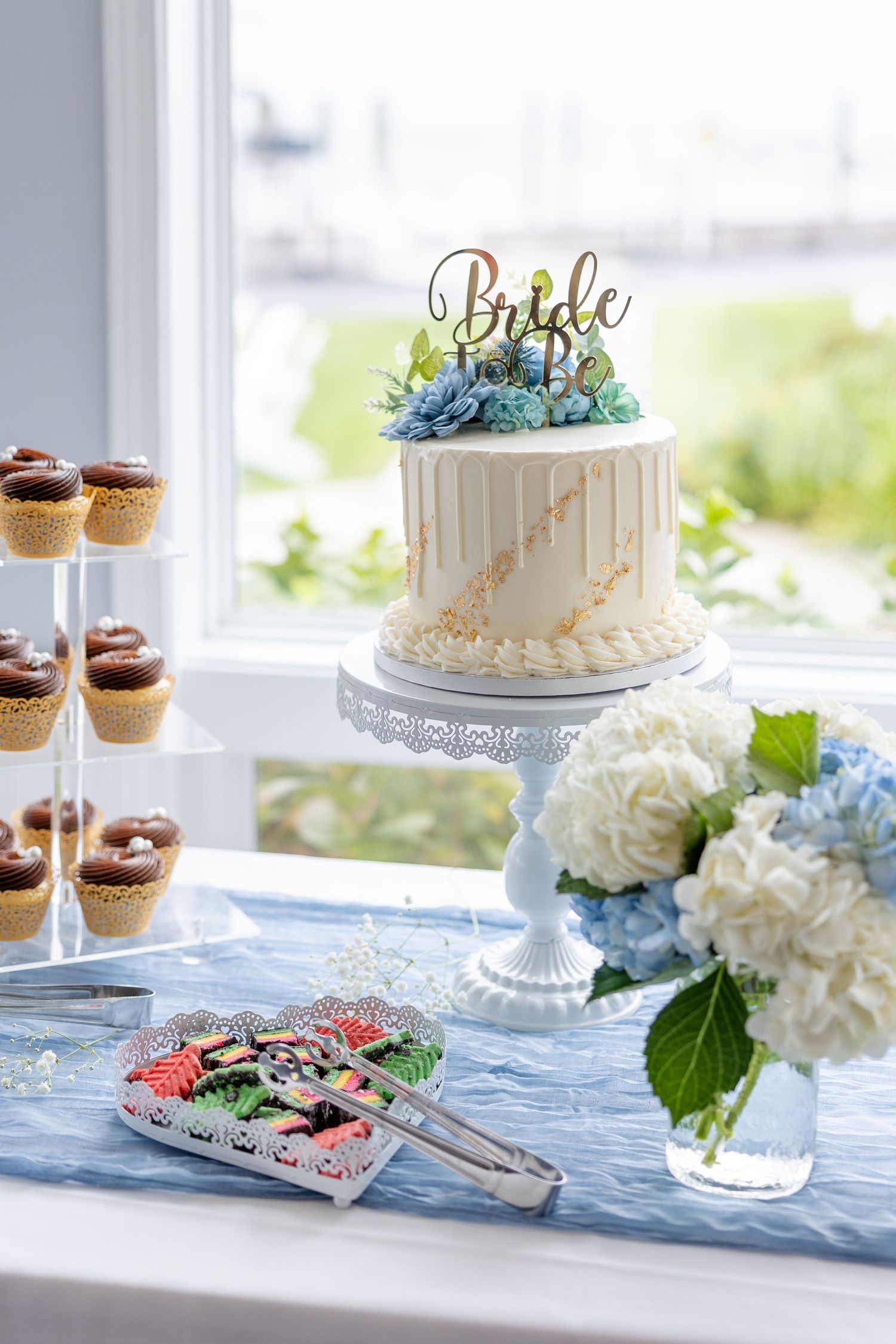 Bridal shower dessert table featuring a white drip cake with Bride topper, cupcakes, and blue hydrangeas.