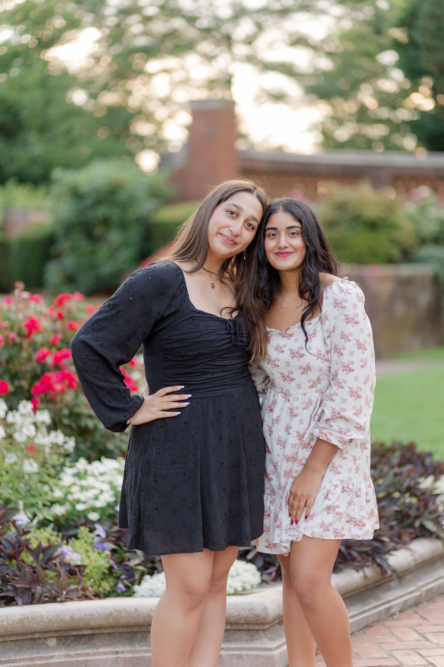 Two young women in floral and black dresses posing together near garden flower bed at sunset.