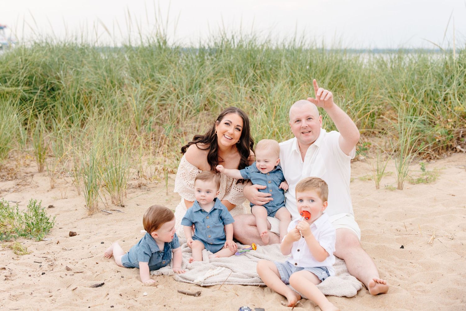 Family with four young boys and grandmother sitting together on sandy beach with tall grass behind them.