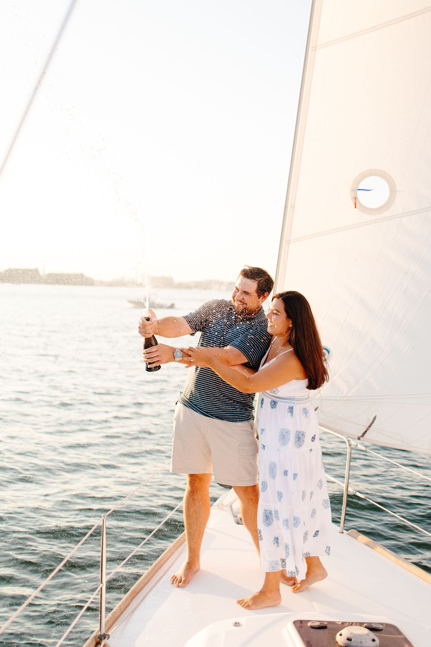 Happy couple celebrating on a sailboat by popping champagne at sunset.