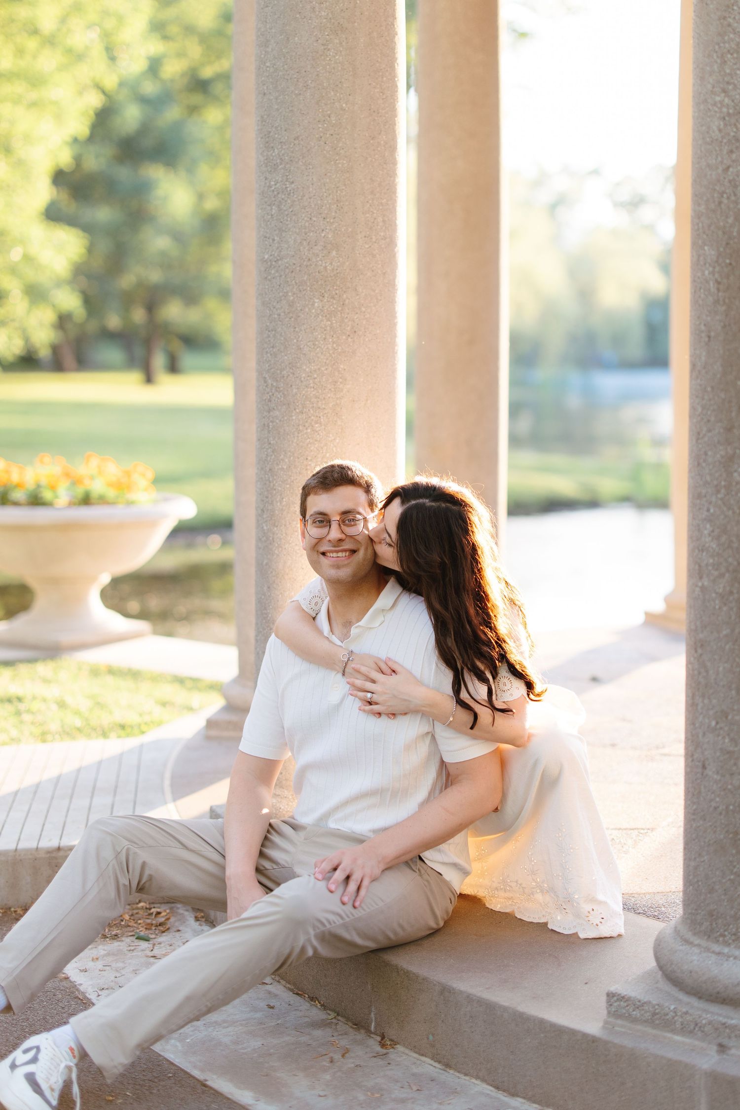 Couple sitting on stone steps between columns in a sunny park, woman kissing mans cheek while he smiles.