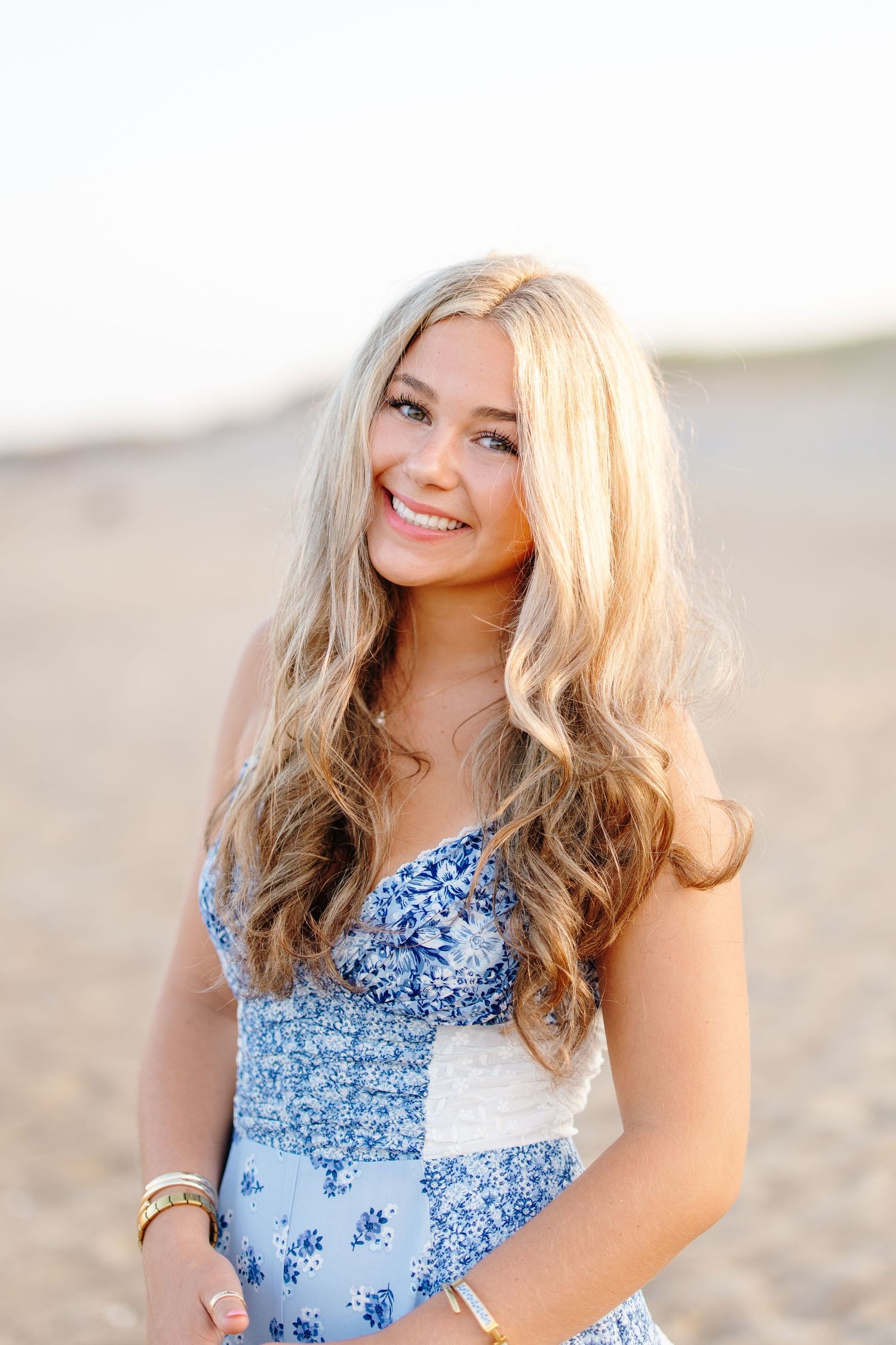 Smiling blonde teen girl in blue floral dress poses on sandy beach during golden hour.