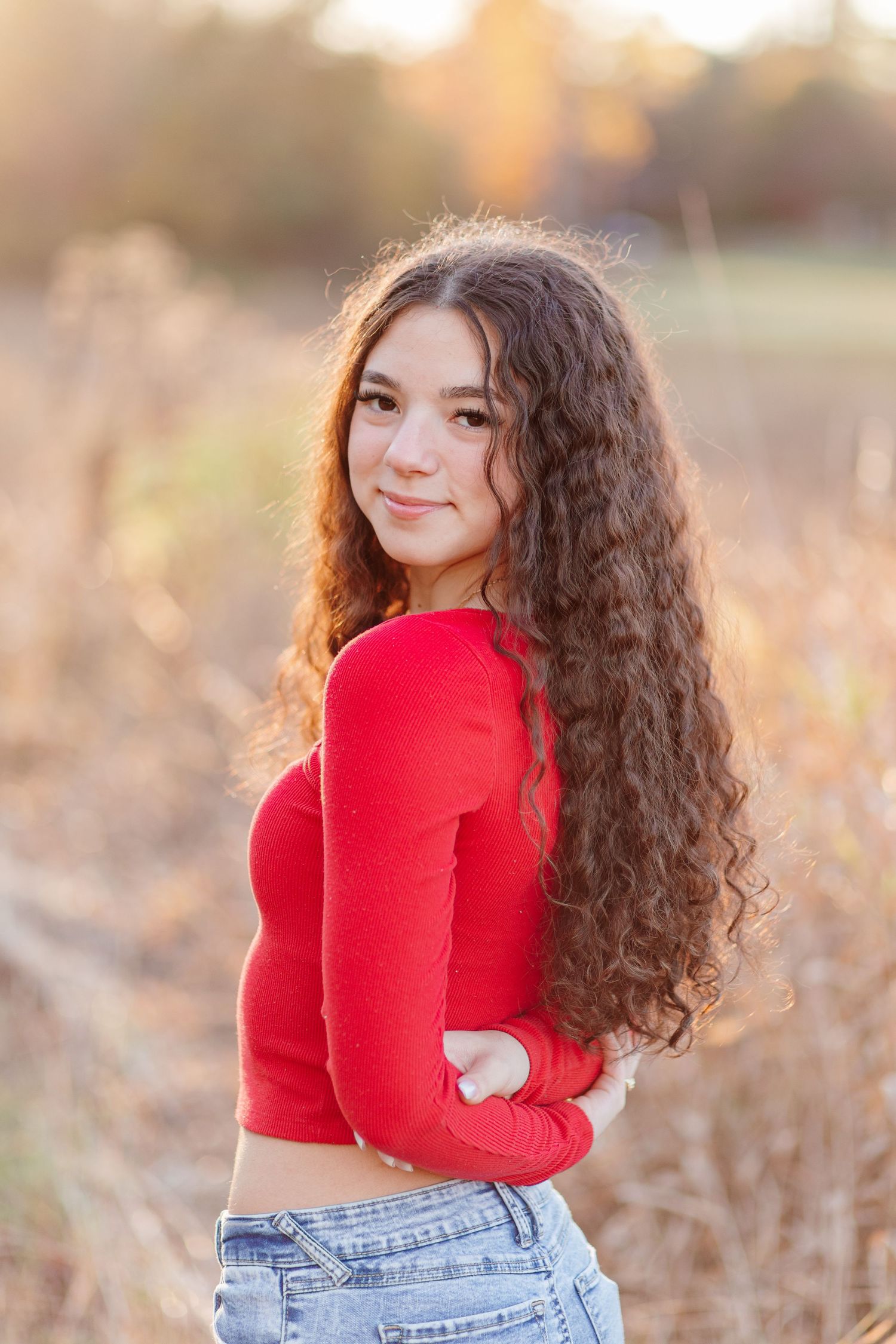 Teen girl with long curly brown hair in red crop top poses in golden field at sunset.