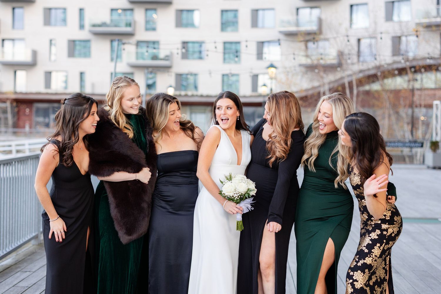 Bride laughing joyfully with six bridesmaids in black and green gowns on outdoor urban walkway.