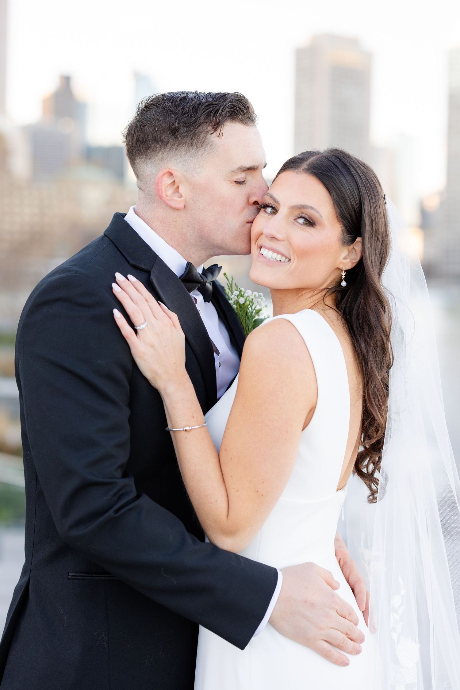 Groom in black tuxedo kisses bride in white gown on cheek with city skyline backdrop.