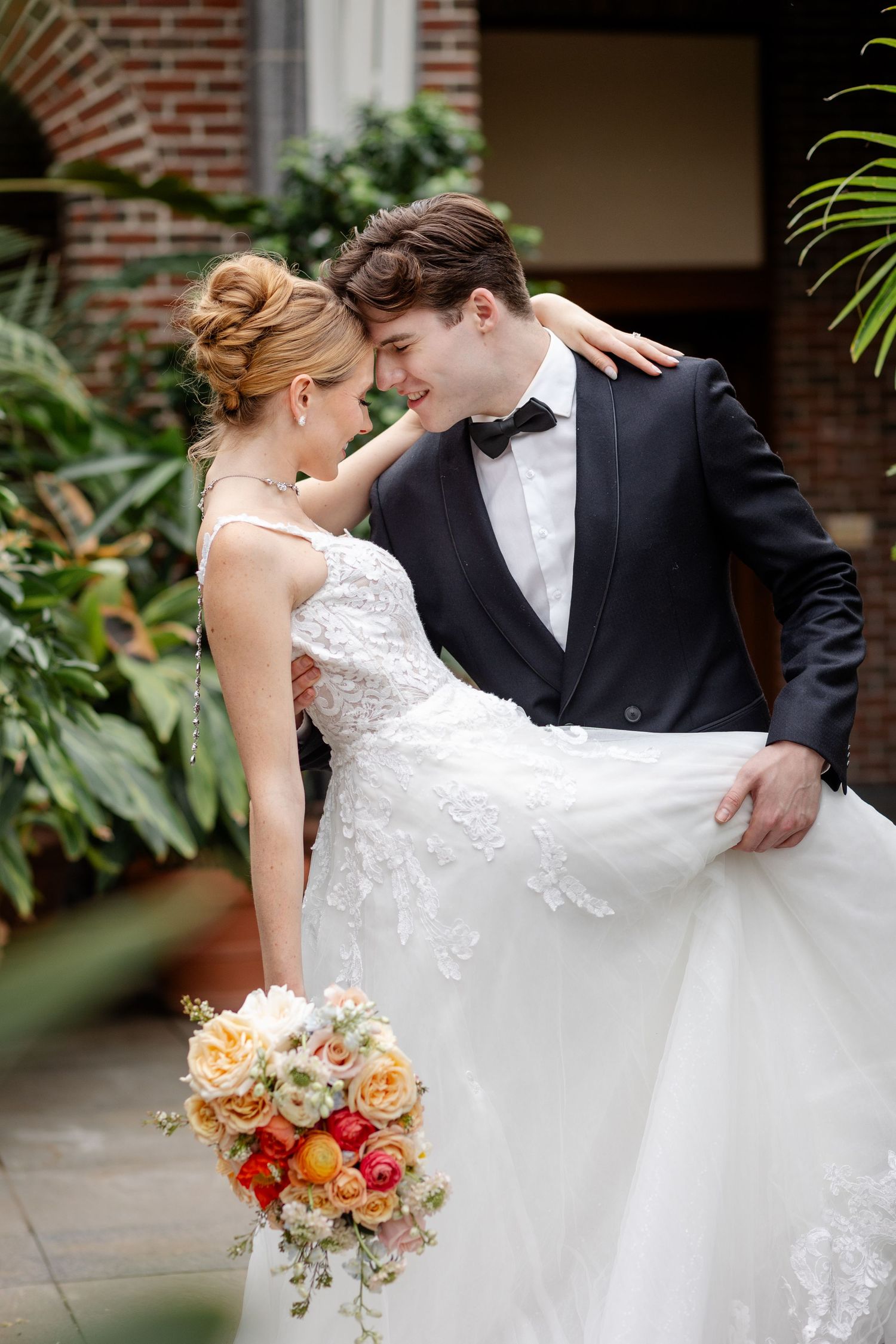Smiling groom lifts bride's gown as couple embraces in green garden setting.