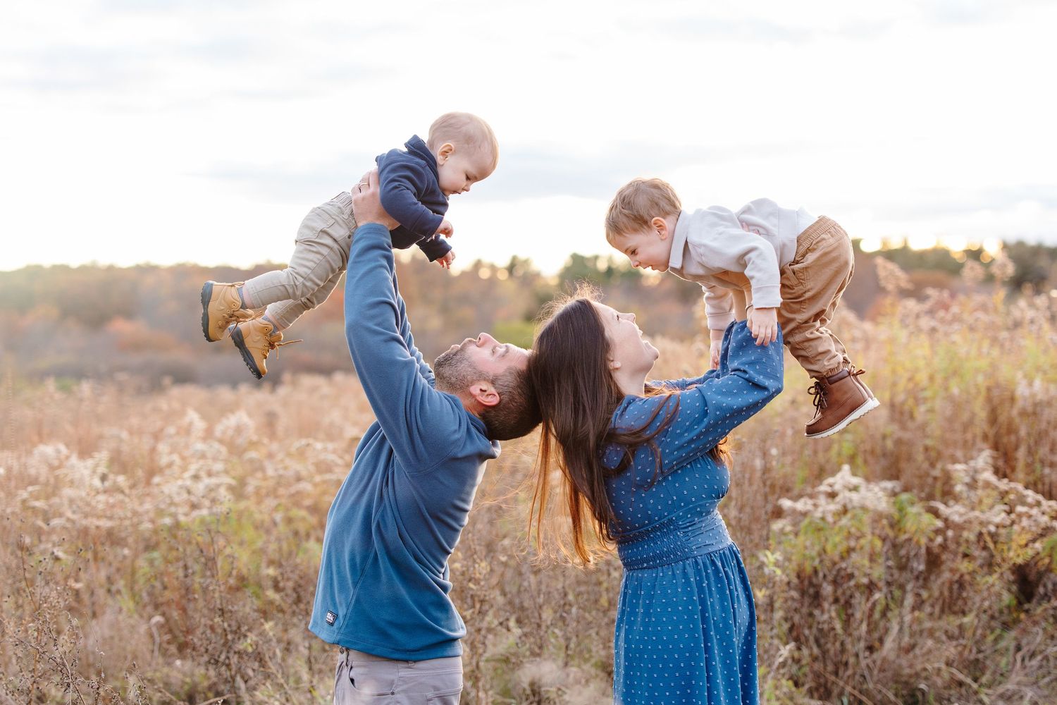 Happy couple lifting two babies in golden autumn field at sunset.
