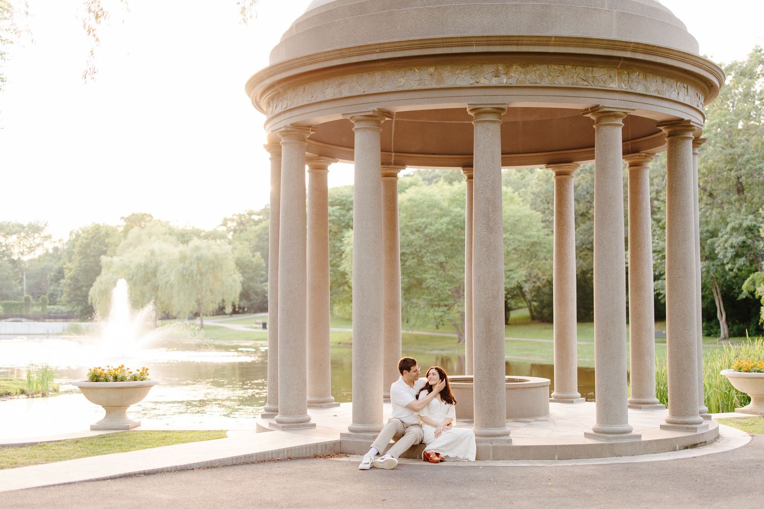 Couple sitting romantically under an elegant white rotunda with columns beside a serene park fountain.