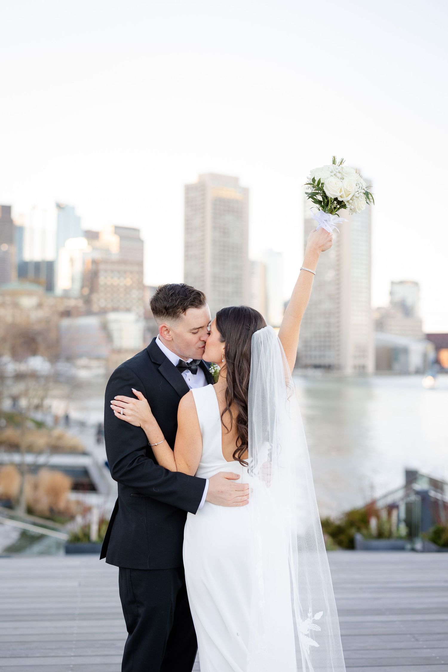 Bride and groom share a kiss on a rooftop with city skyline backdrop, bride raising bouquet in celebration.