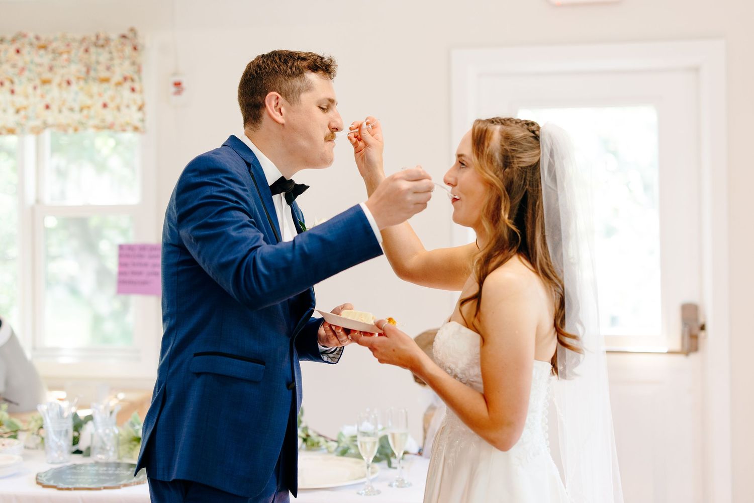 Groom in blue suit feeds cake to smiling bride in white dress during fun wedding reception moment.