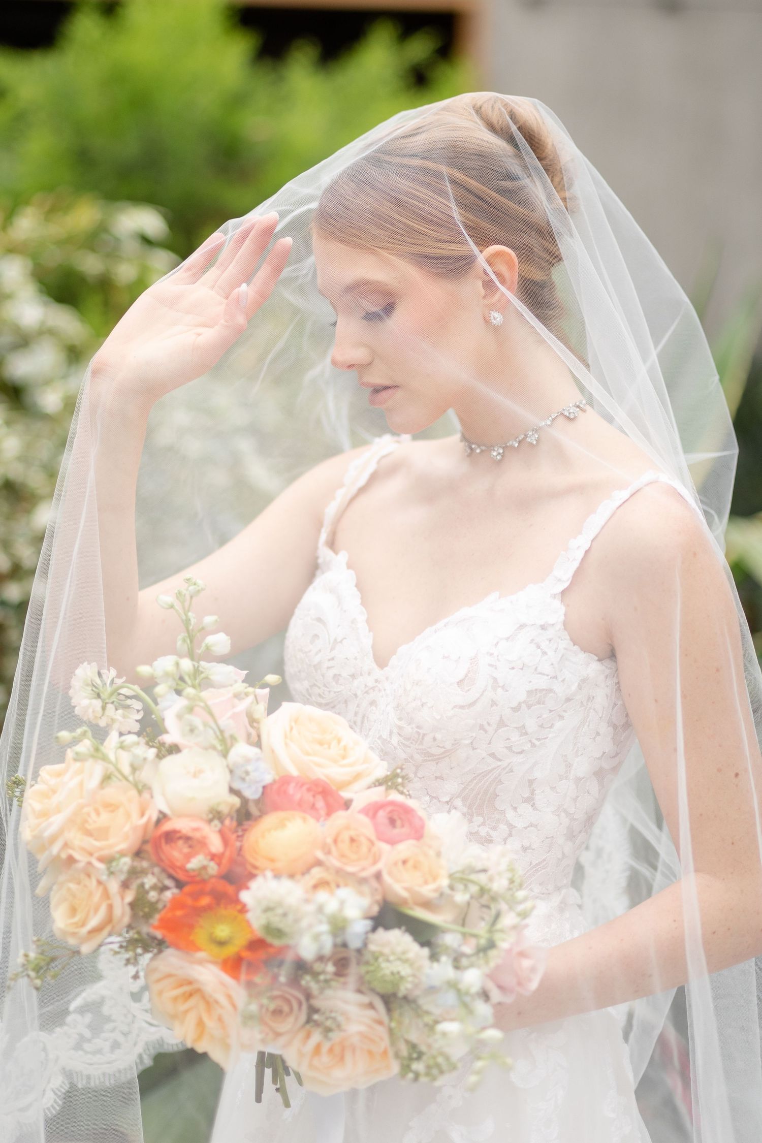 Bride in lace gown holds colorful bouquet under sheer veil outdoors.