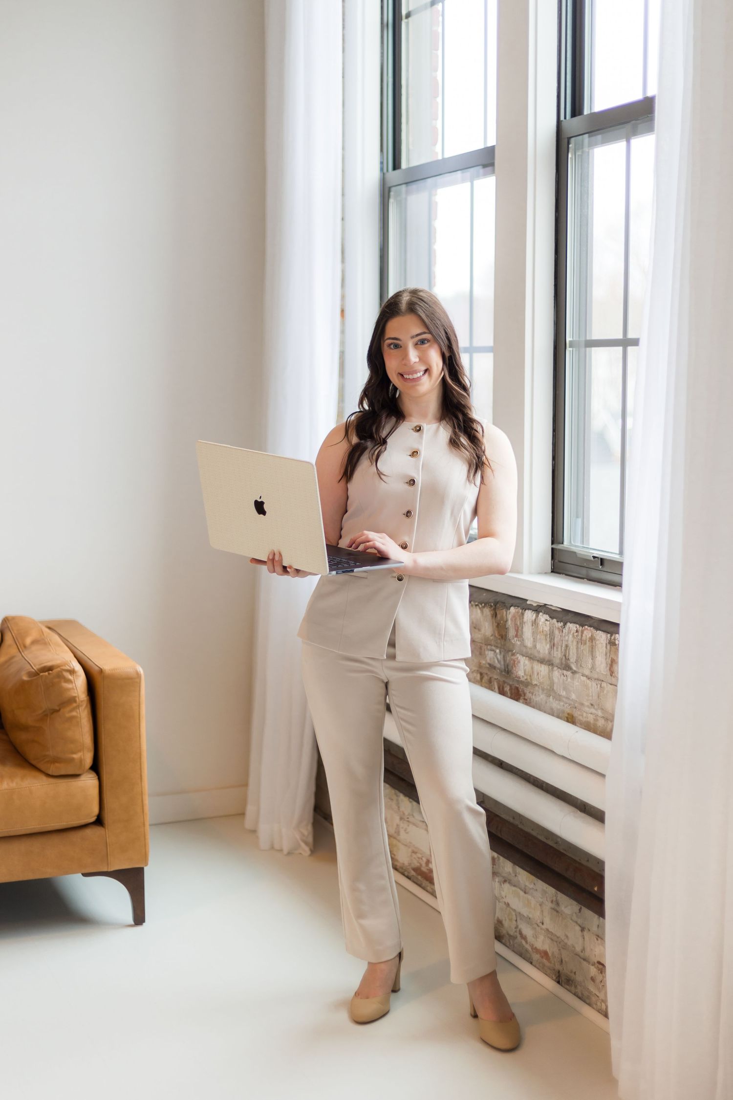 Smiling businesswoman in beige outfit holding Apple MacBook laptop while standing near bright windows.