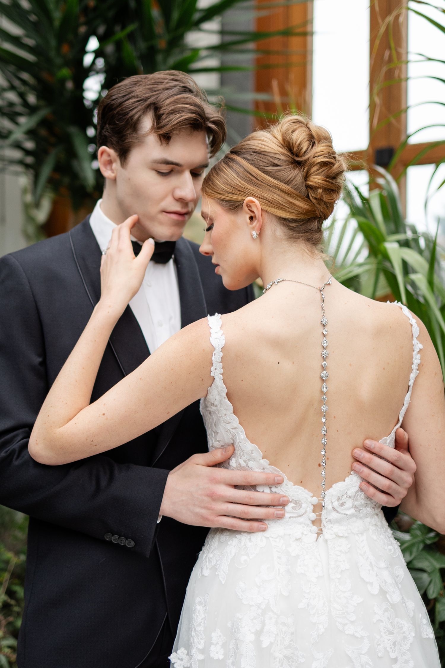 Groom in black tuxedo embraces bride showing stunning open back lace wedding gown.
