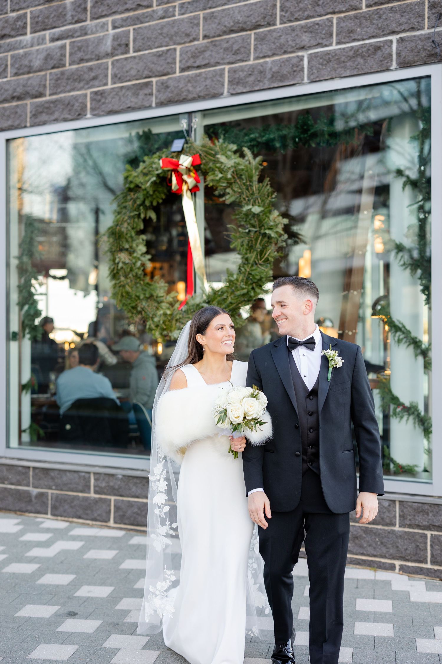 Bride and groom pose outside a building adorned with a Christmas wreath, bride holding white bouquet in winter wedding attire.