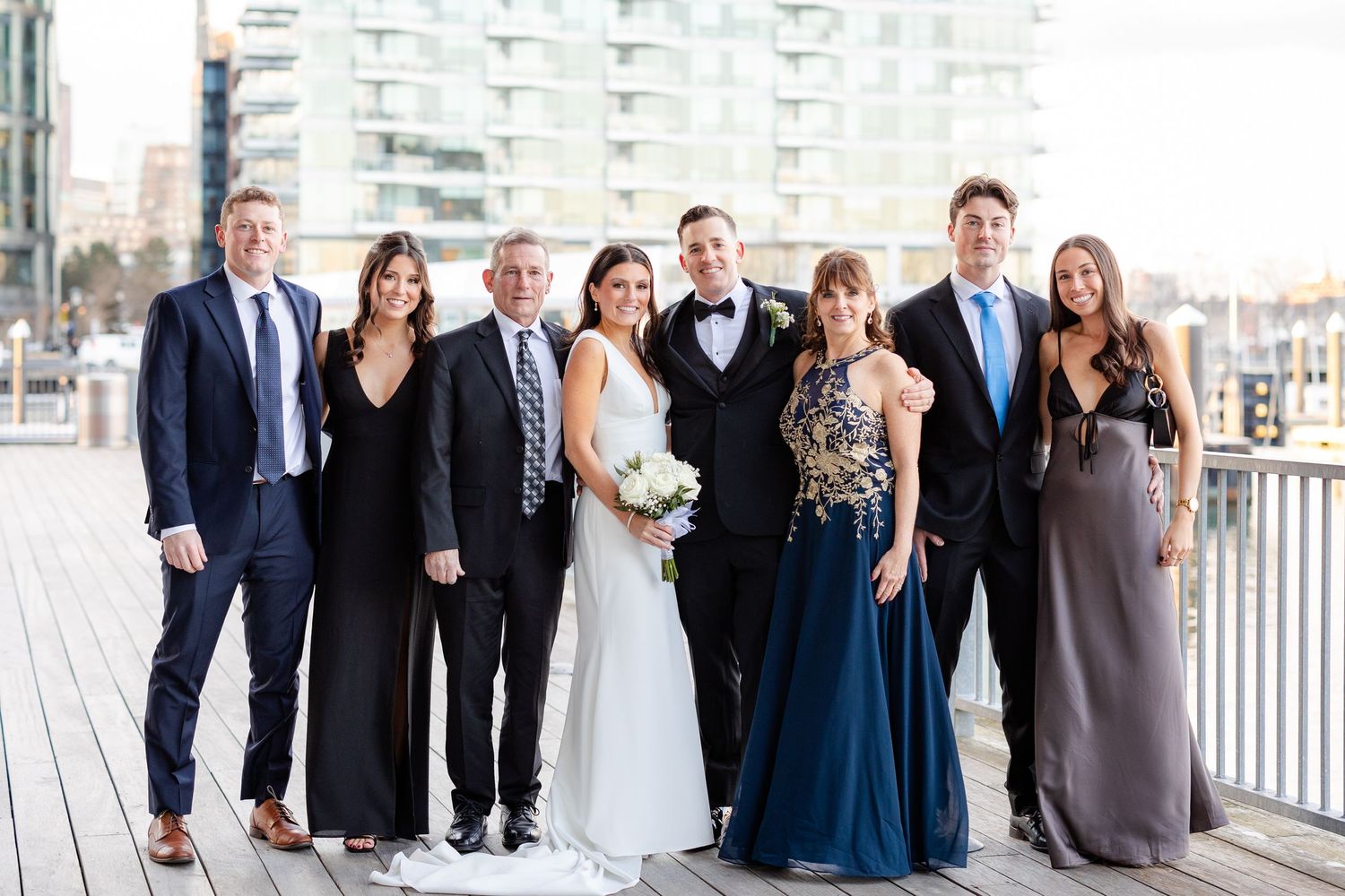 Wedding party and family pose together smiling on waterfront dock with city harbor views behind them.