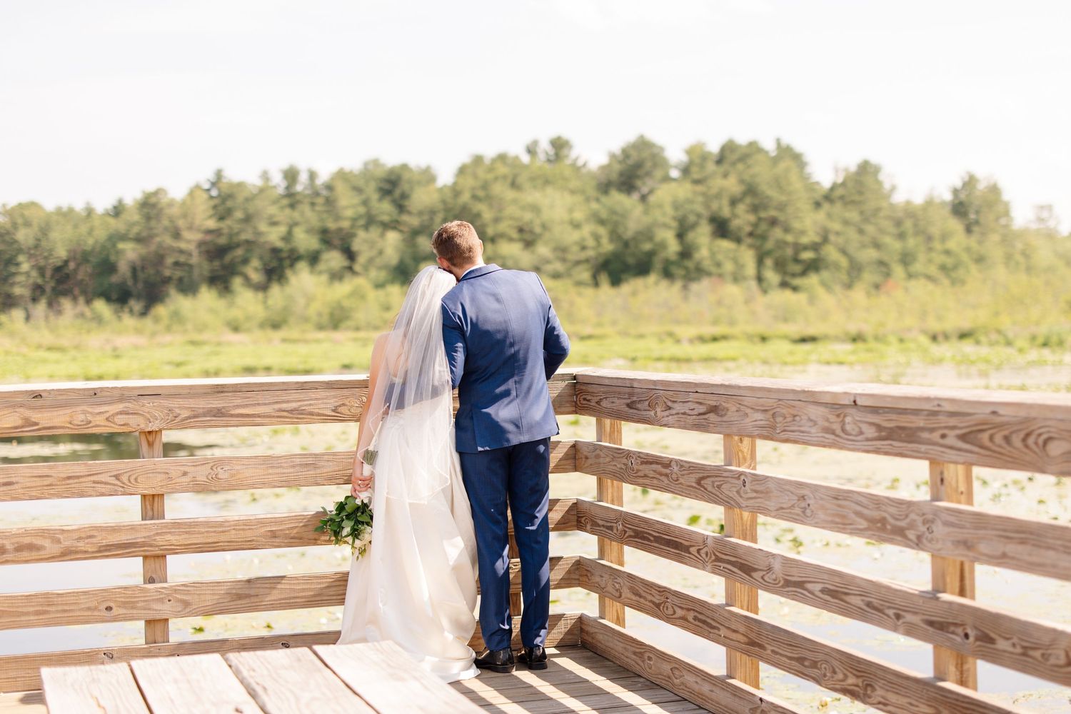 Bride and groom stand together facing a scenic wetland view from a wooden deck on their wedding day.