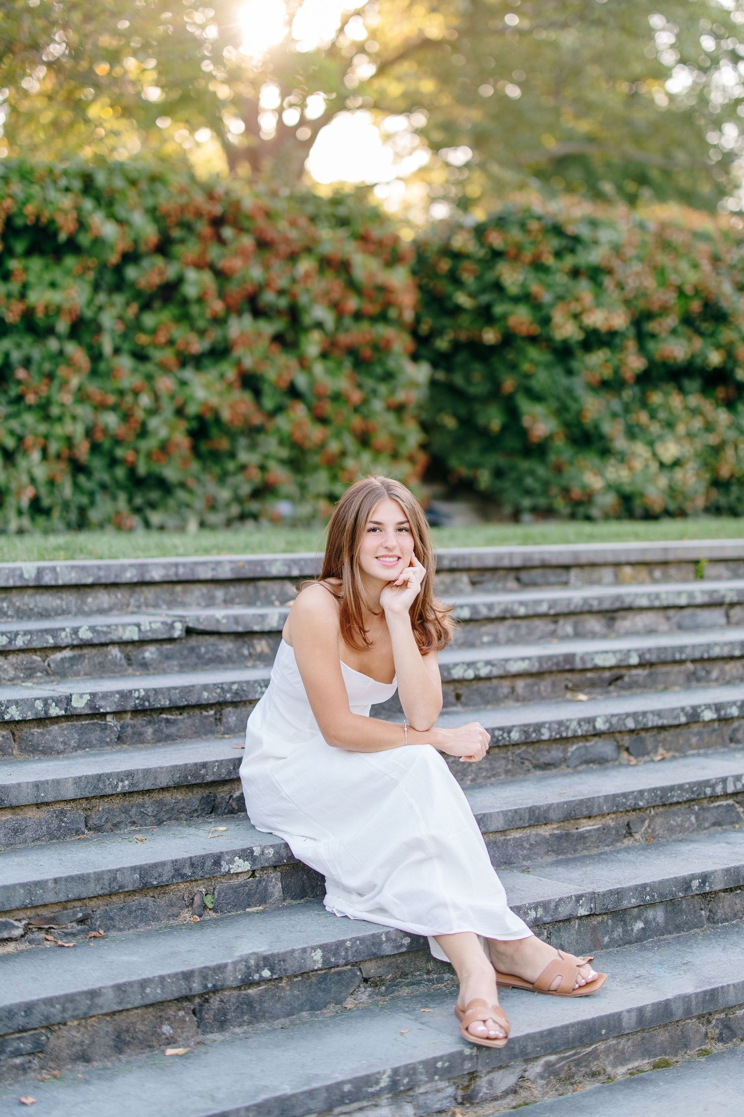 Smiling teen girl in white dress sits on stone steps with lush green garden backdrop.