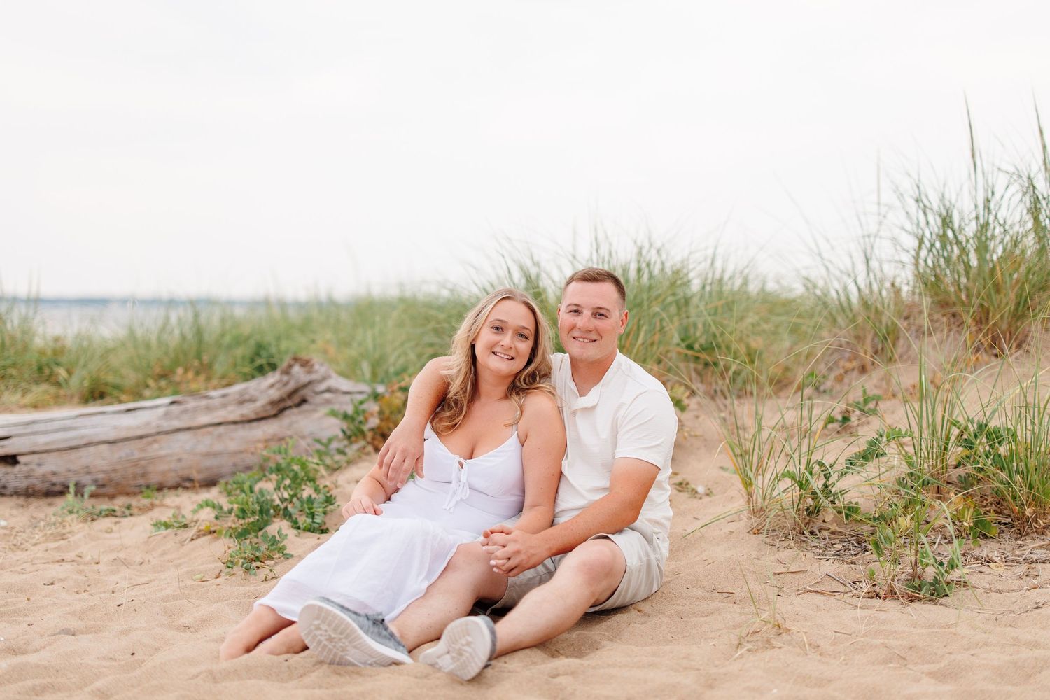 A smiling couple sits together on sandy beach dunes surrounded by tall grass during an outdoor photo session.