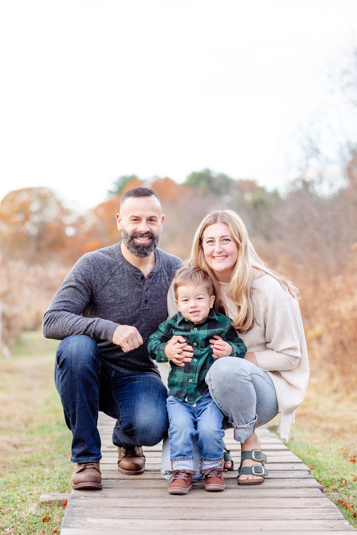 A happy family of three poses on a wooden boardwalk surrounded by autumn foliage during an outdoor portrait session.