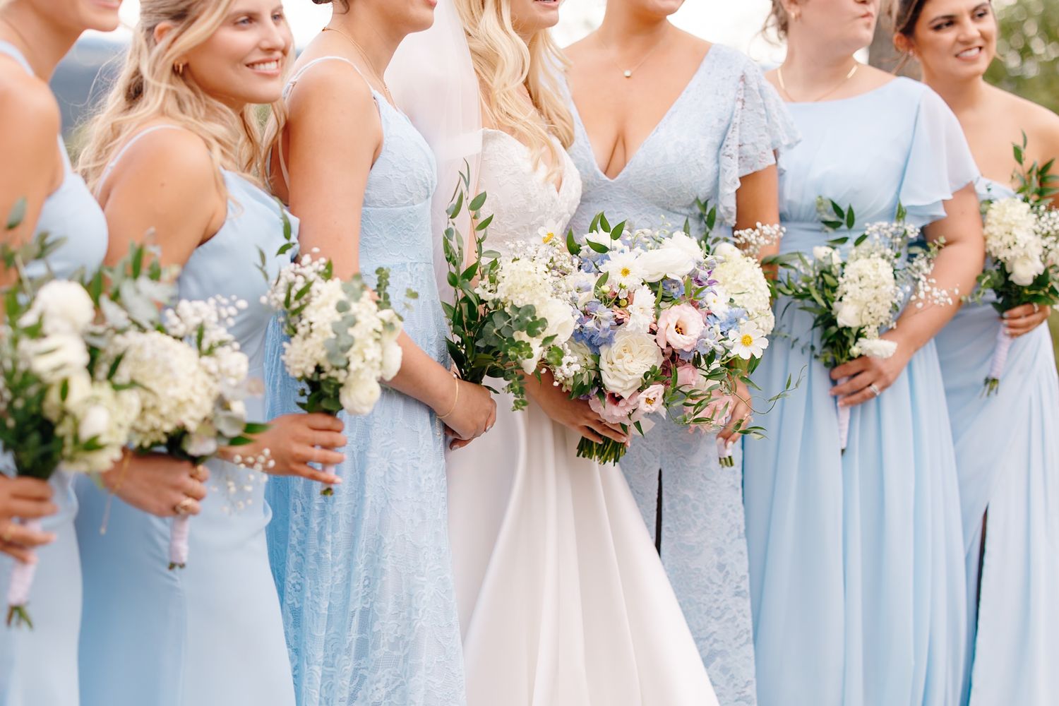 Bridesmaids in light blue dresses holding white and pastel floral bouquets alongside a bride in white.