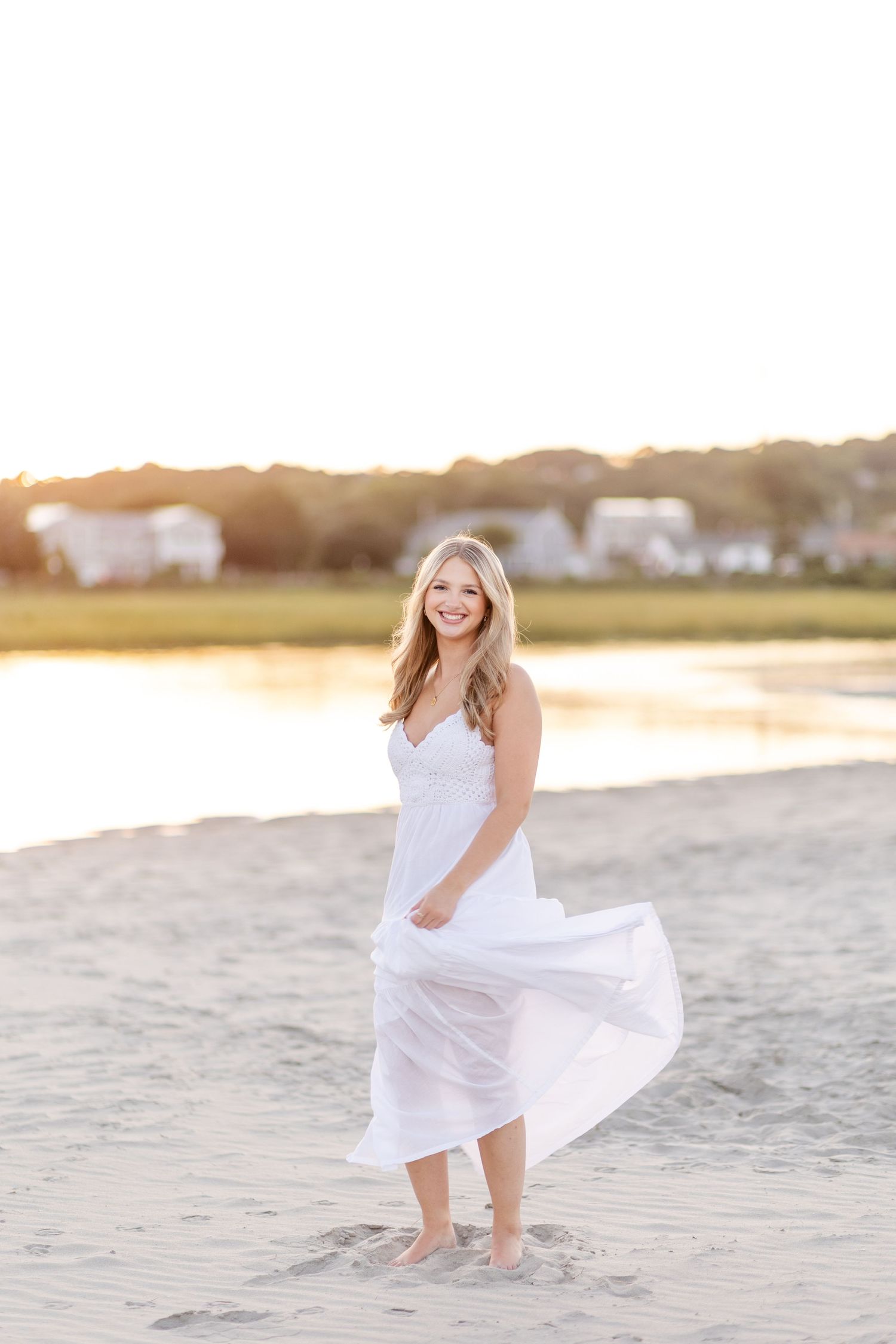 Blonde woman in flowing white dress twirling on a sandy beach at sunset.