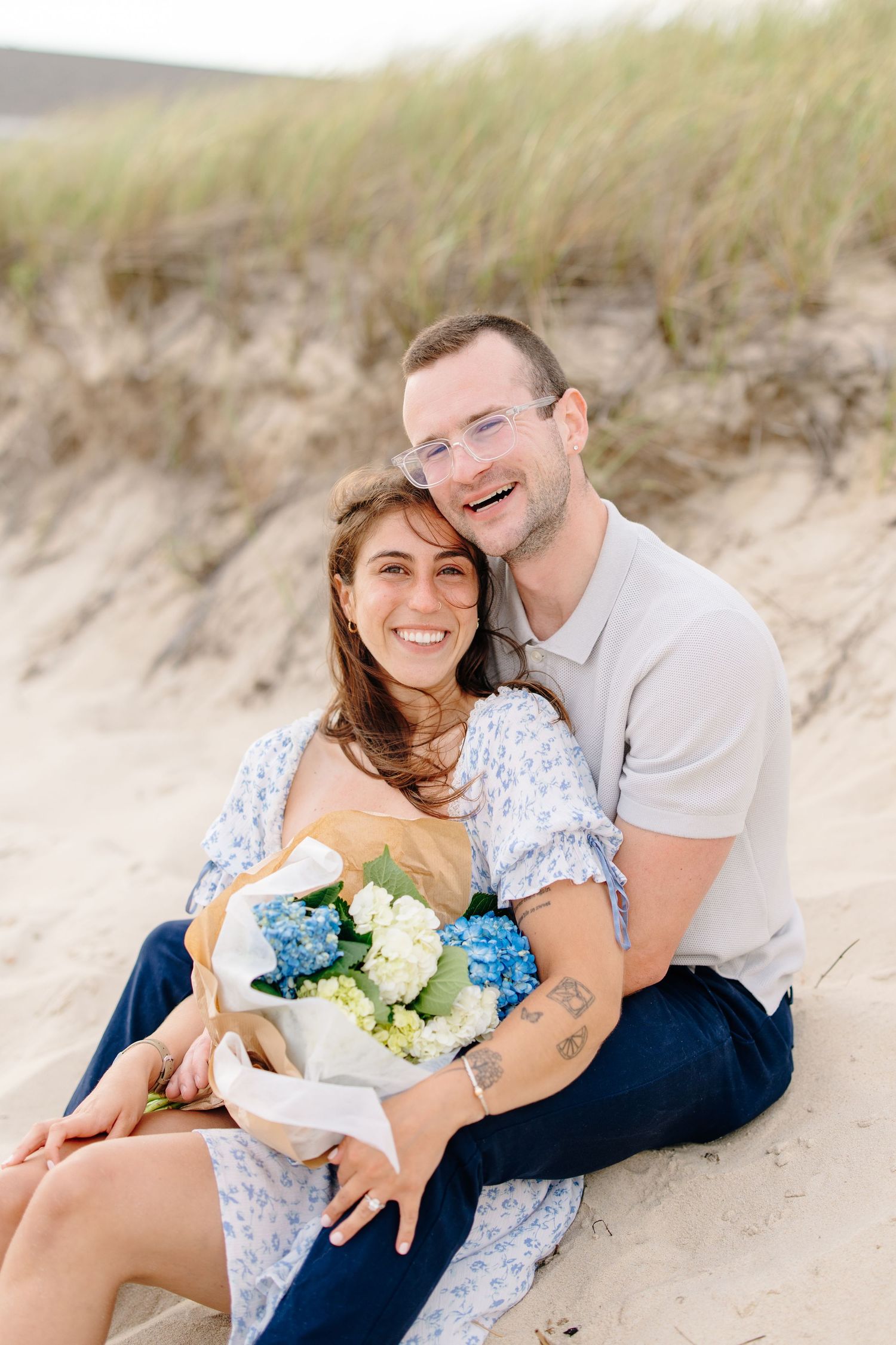 Smiling engaged couple sitting on sand dunes holding a bouquet of blue and white hydrangeas.