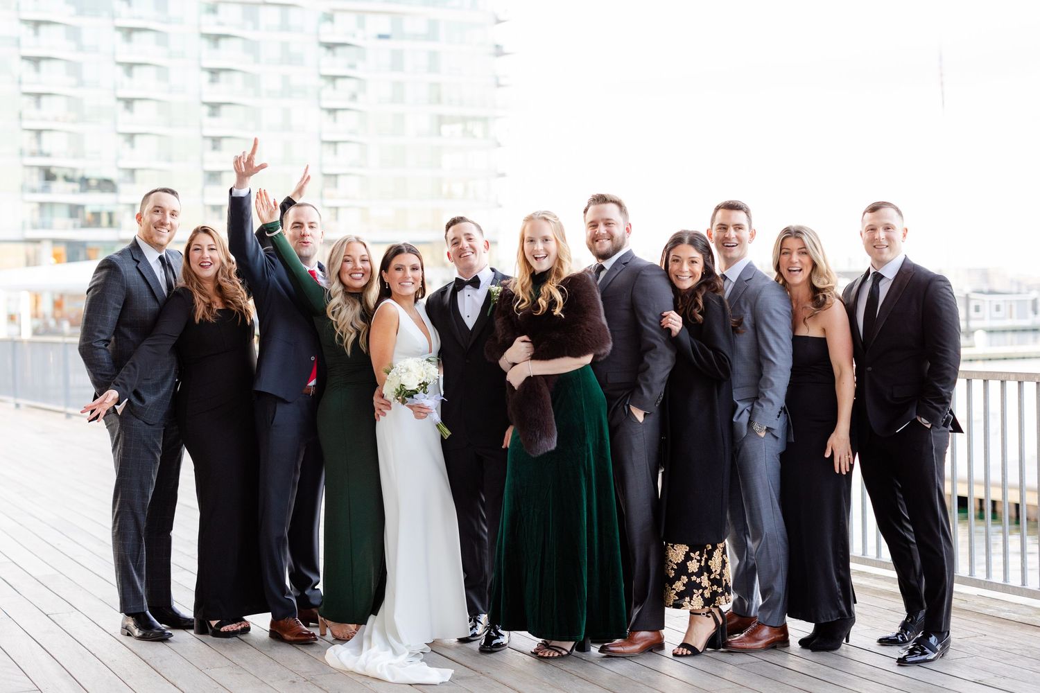 Wedding party of ten poses together smiling on wooden deck with urban building in background.