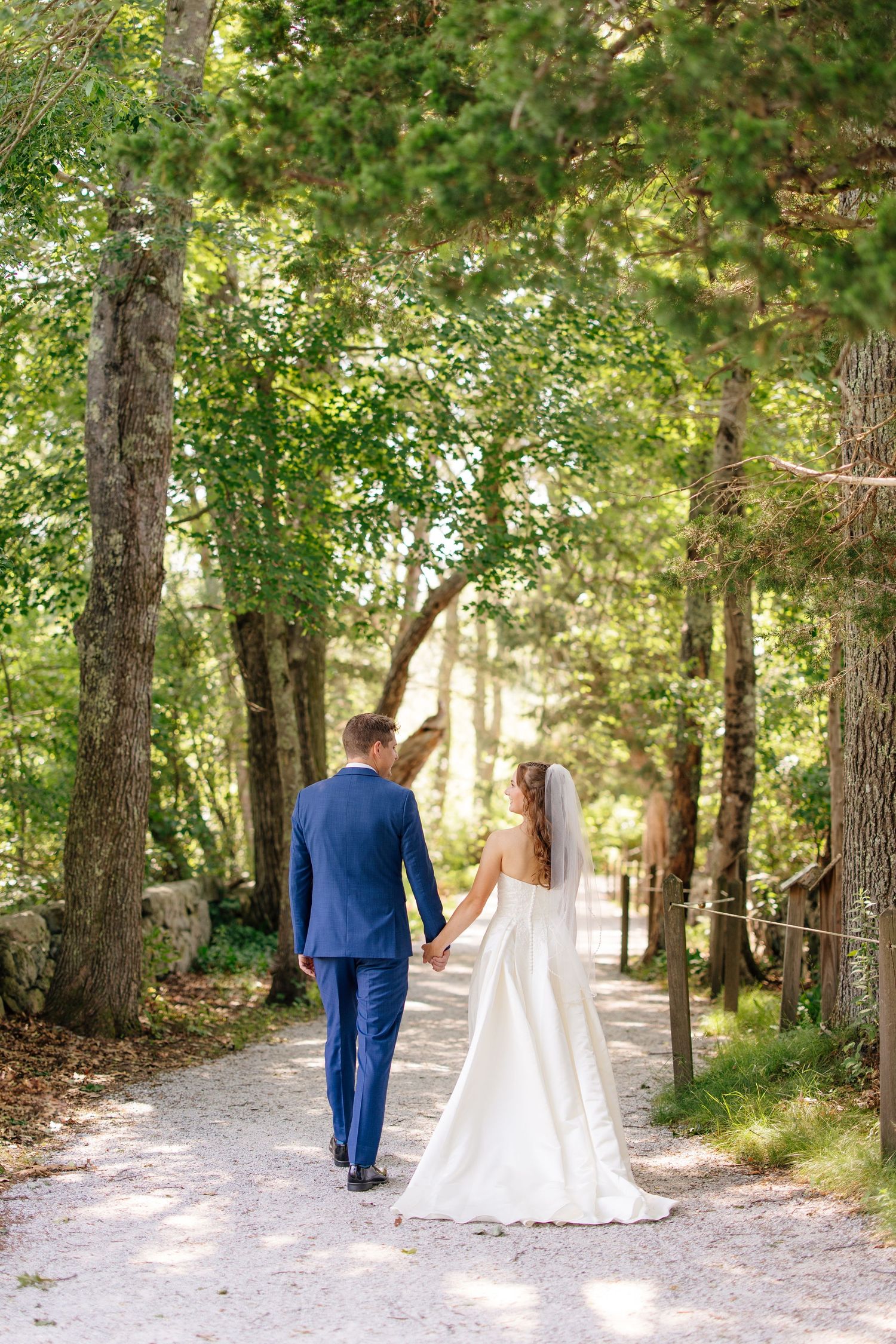 Bride and groom walking hand in hand down tree-lined path at outdoor wedding venue.