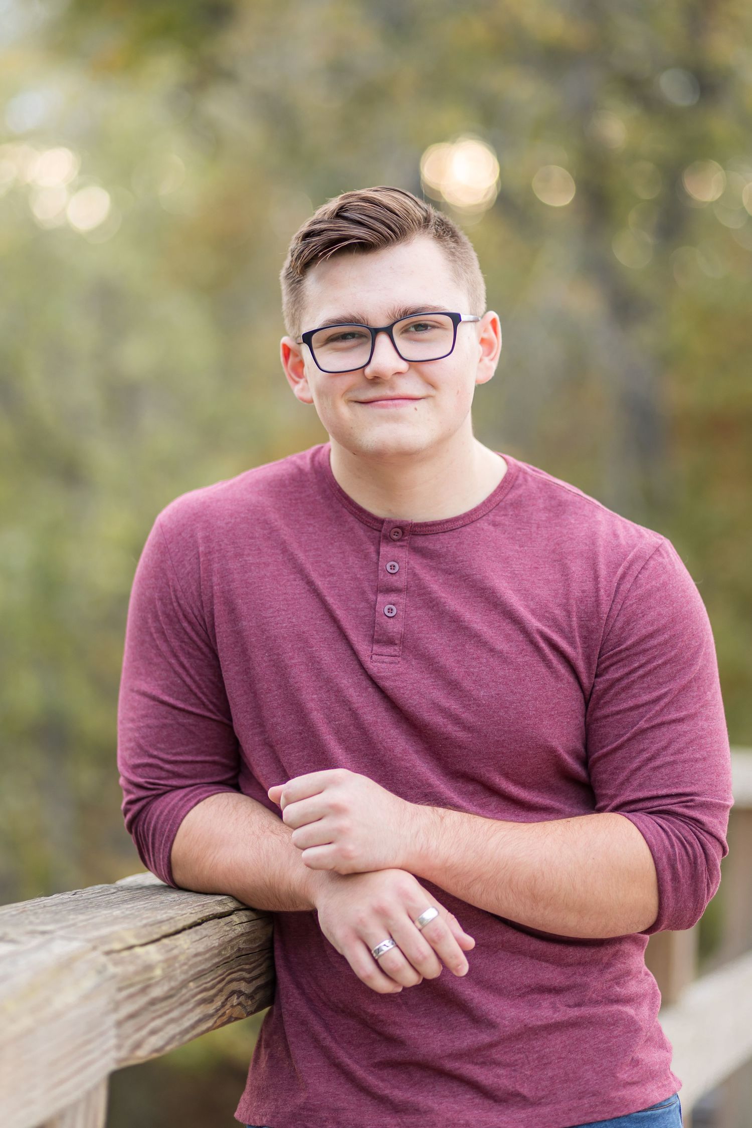 A young man in a maroon henley shirt leaning against a wooden railing outdoors, wearing glasses and smiling.