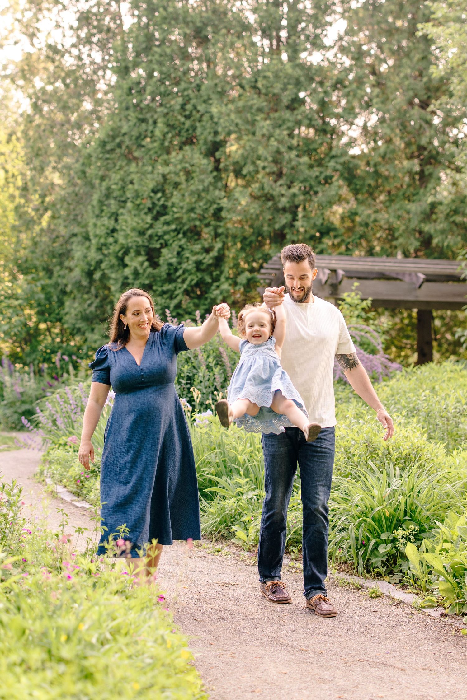 A smiling family of three walks along a garden path, parents swinging their toddler daughter between them.