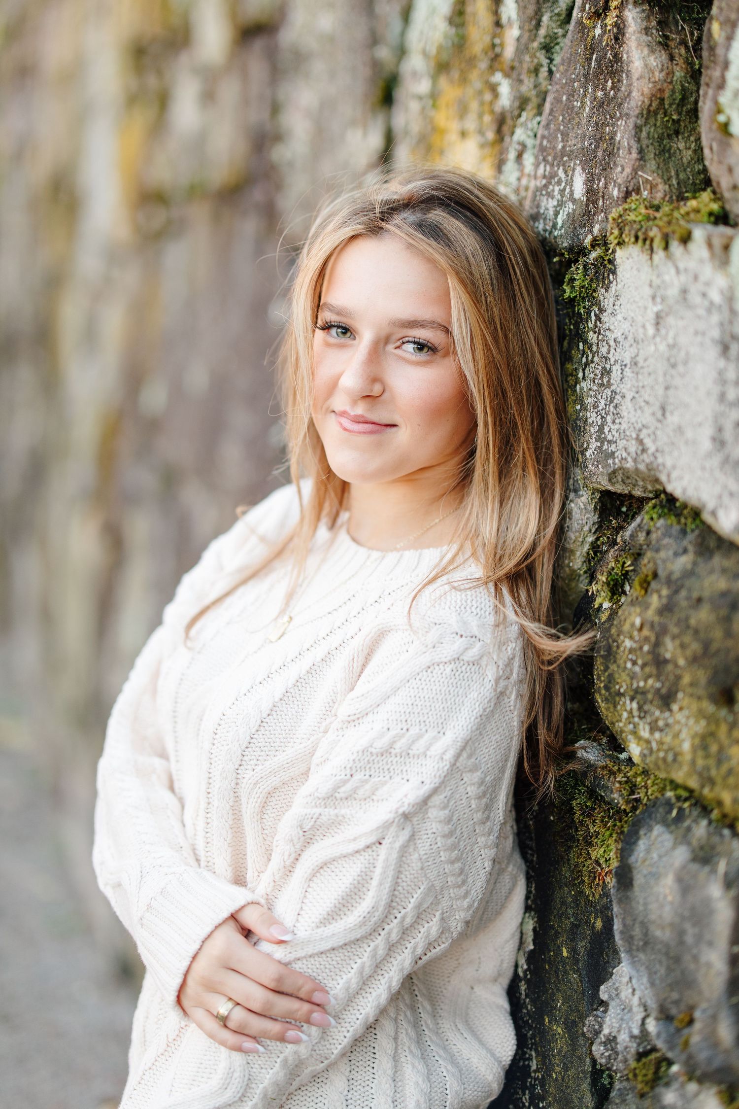 Teen girl with blonde hair in white cable knit sweater leans against mossy stone wall.