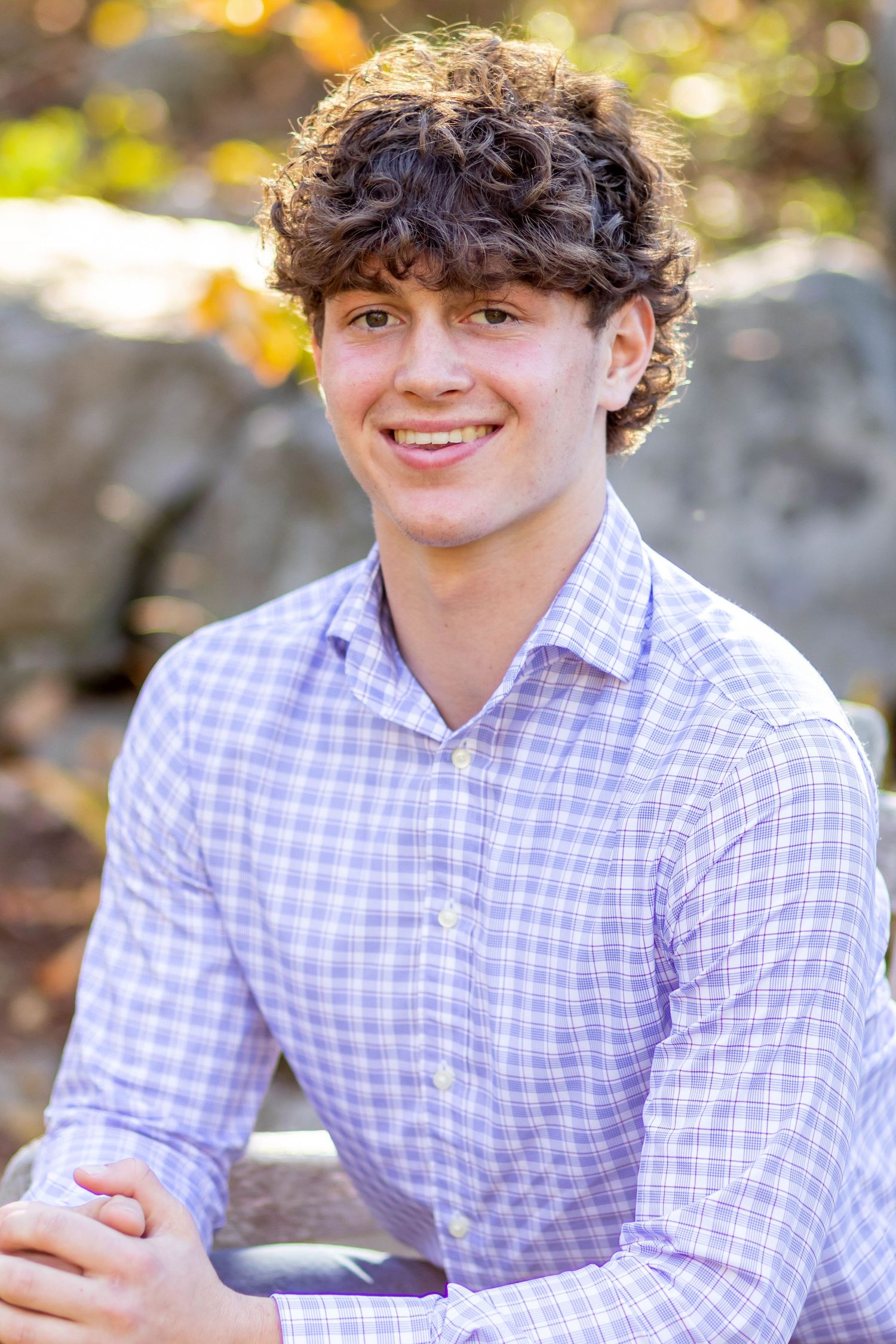 A smiling young man with curly hair wearing a light blue checkered shirt, seated outdoors with fall foliage.