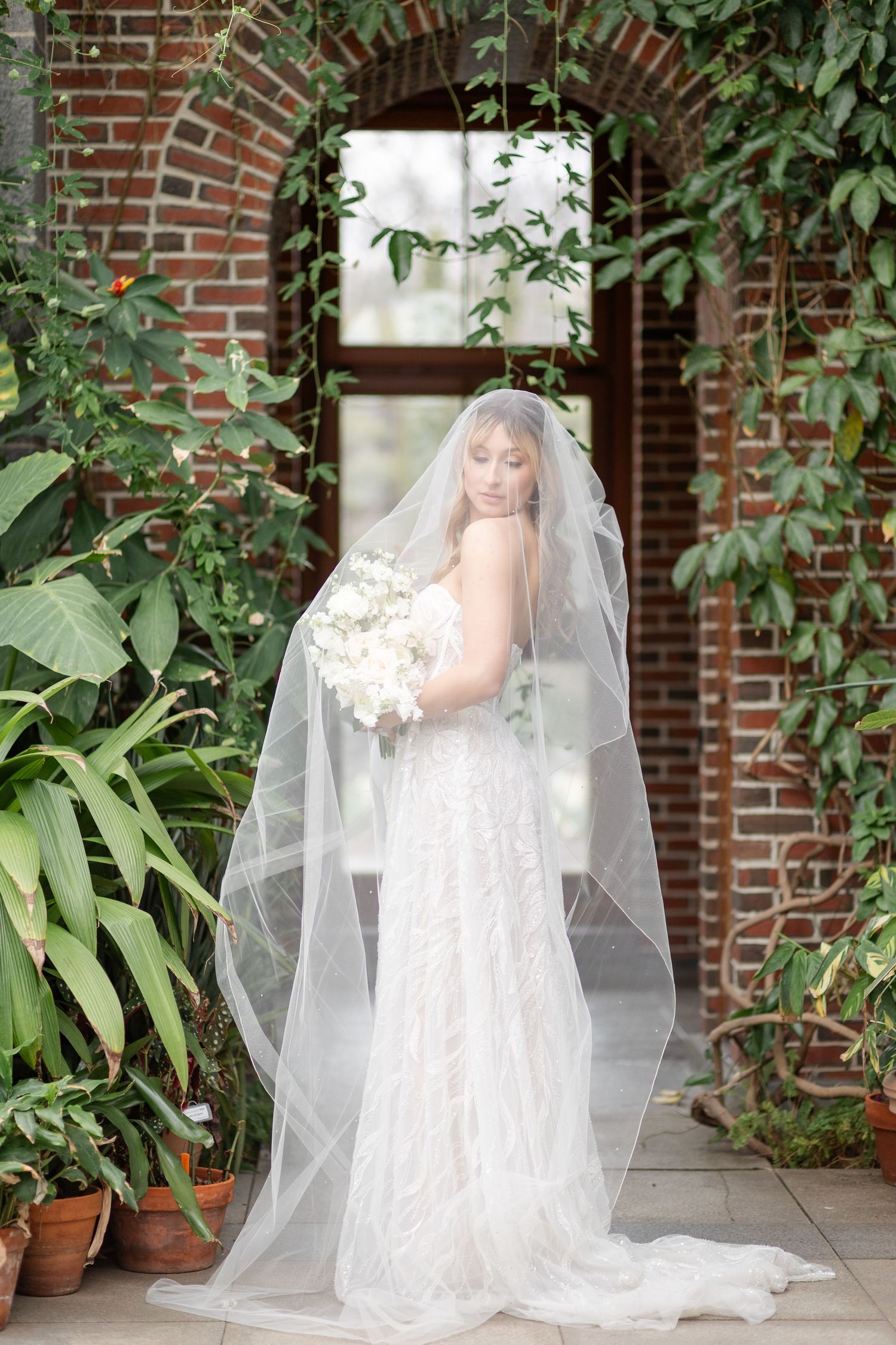 Bride in lace gown and cathedral veil holds white bouquet before ivy-covered brick archway.