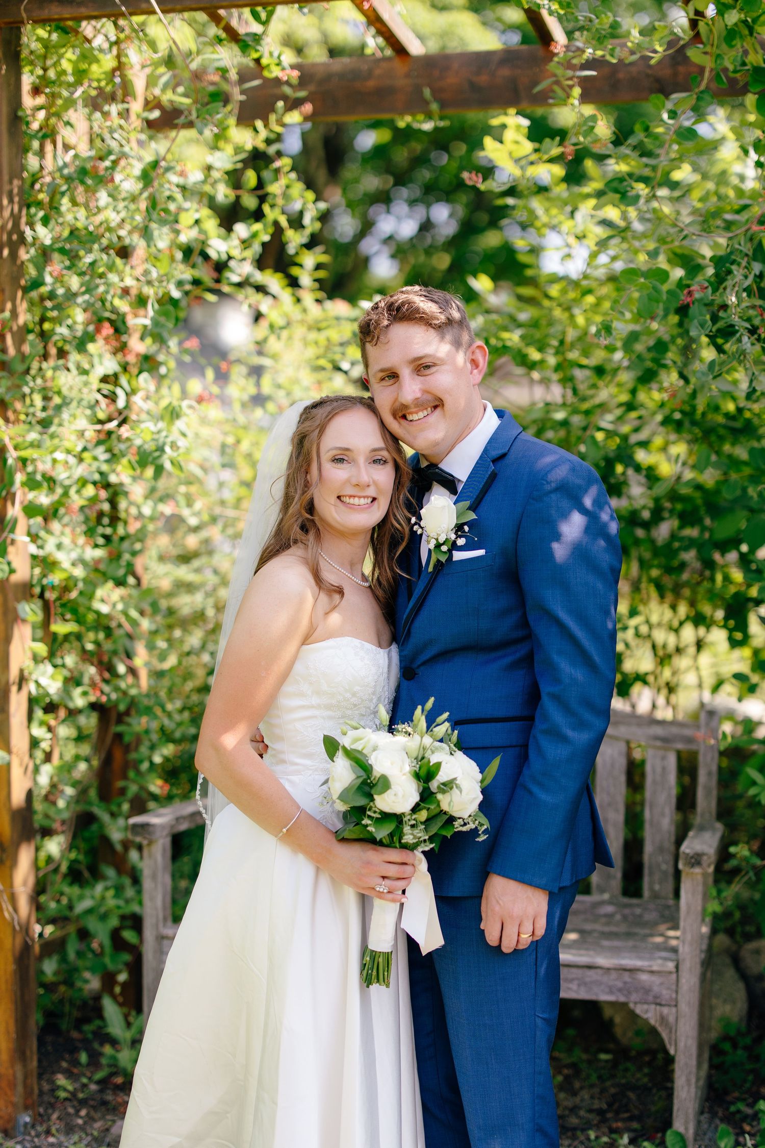 Bride and groom smiling under wooden pergola with lush greenery, holding white floral bouquet.