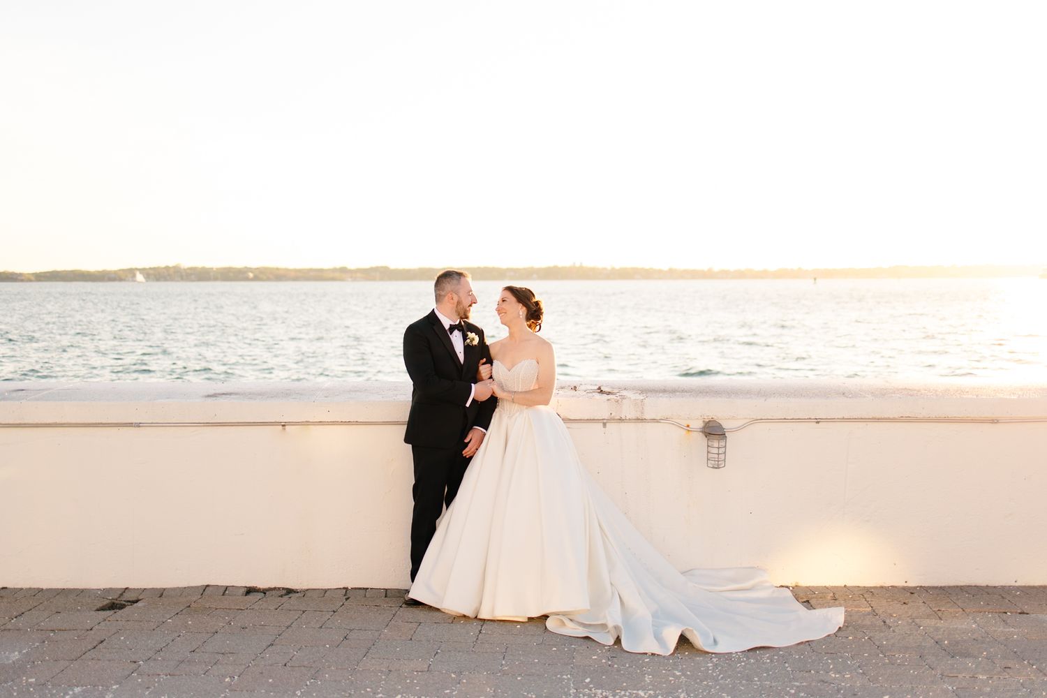 Bride and groom sharing a tender moment leaning against a waterfront wall at golden hour sunset.