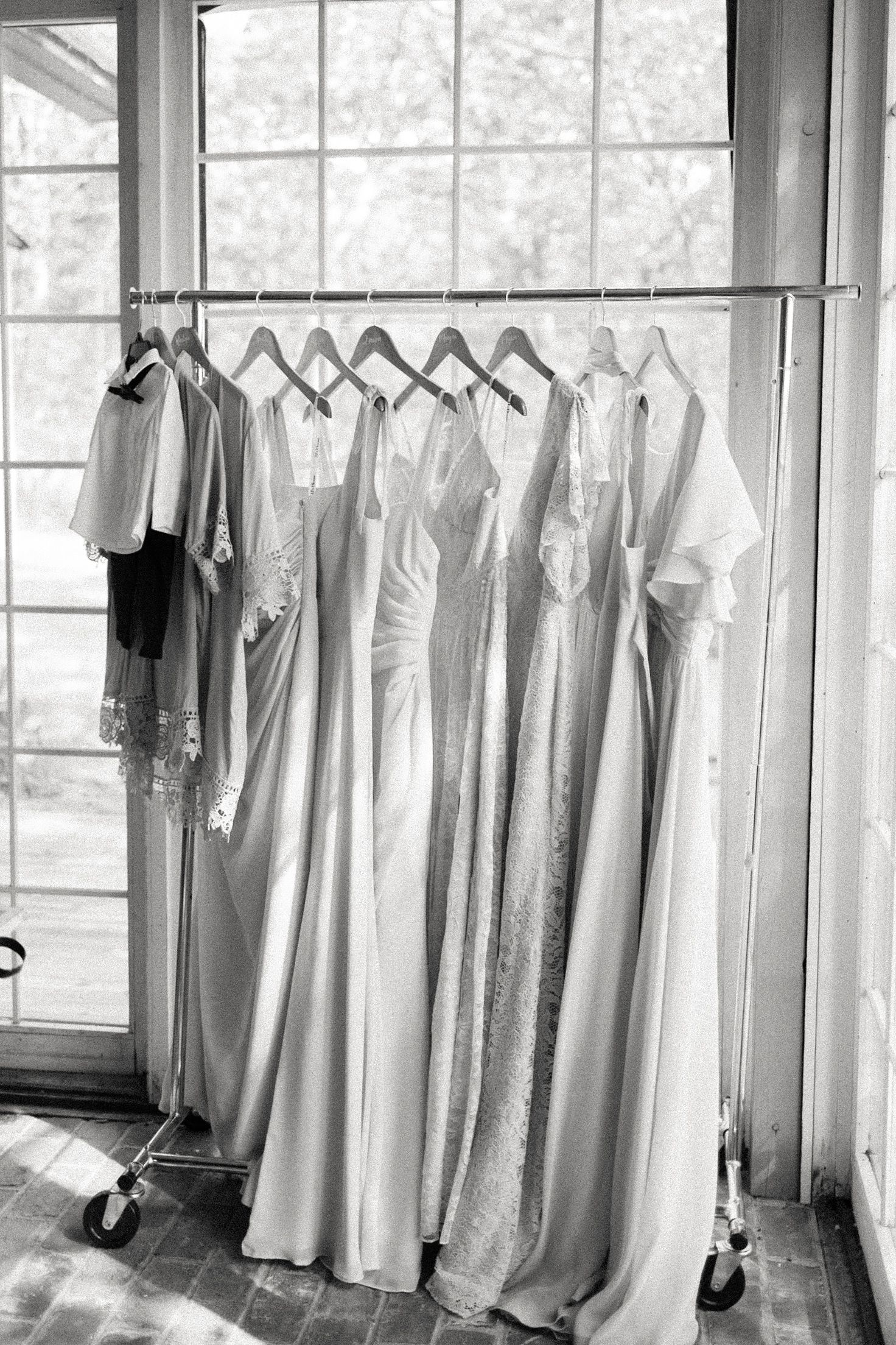 Black and white photo of elegant bridesmaid dresses and bridal gown hanging on a rolling rack near a window.
