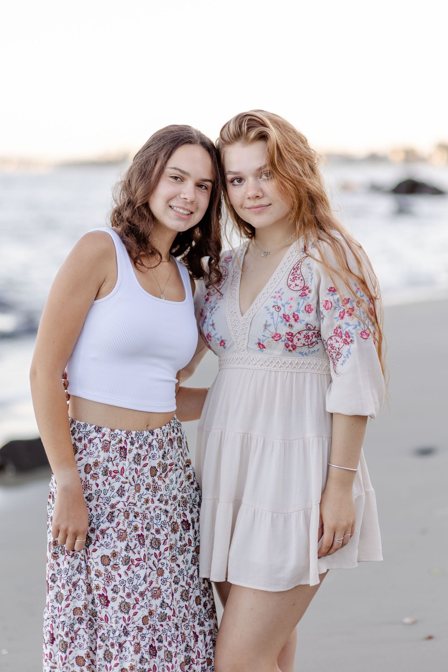 Two teenage girls smiling together on a beach, one in a white crop top and floral skirt, the other in a floral dress.