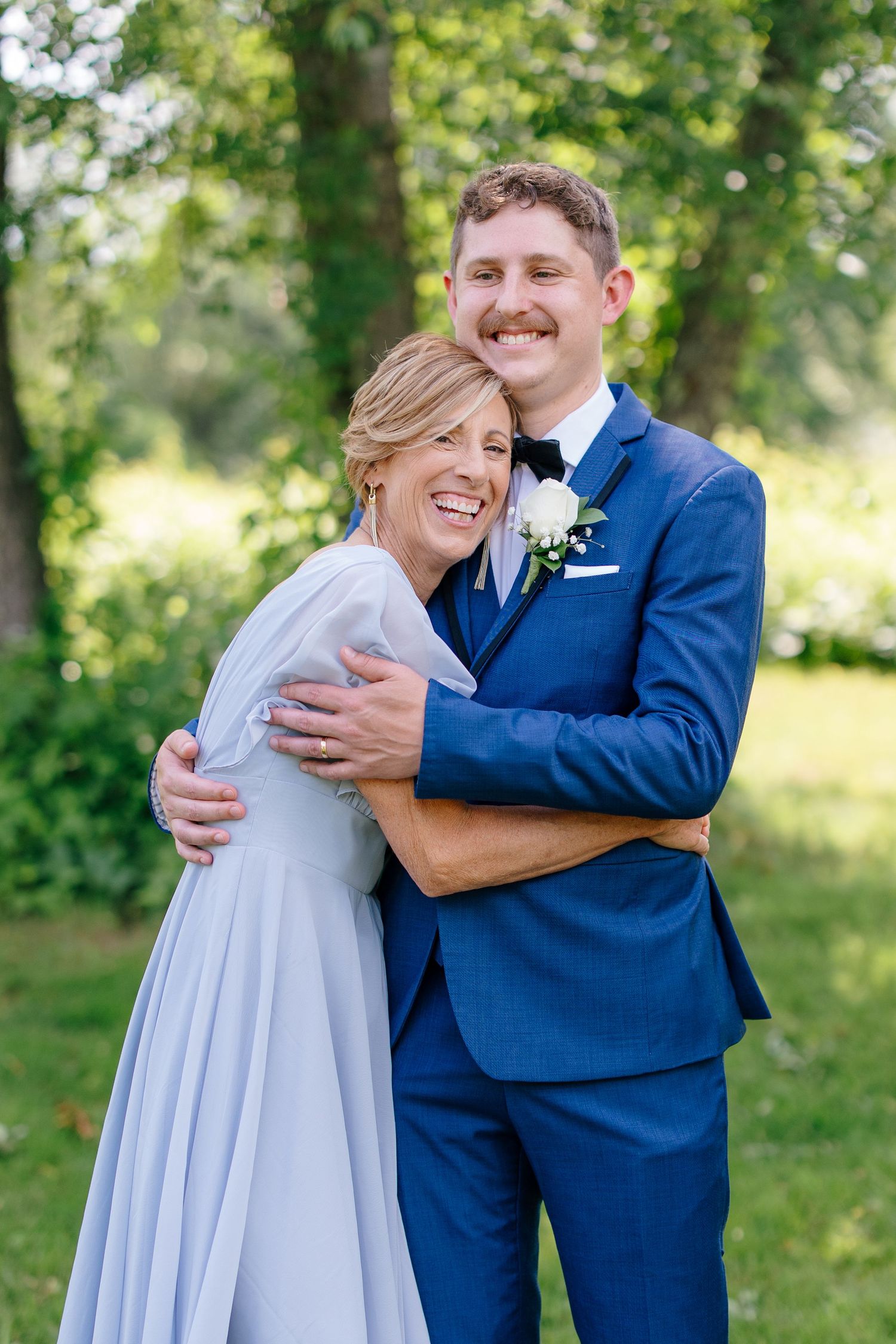 Groom in blue suit embracing smiling woman in lavender dress at outdoor wedding celebration.