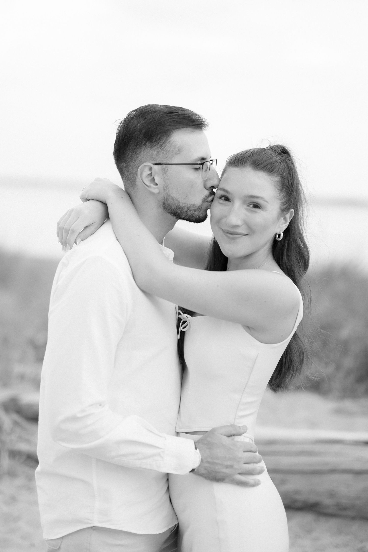 Black and white photo of man kissing woman on cheek as she smiles, beach grass in background.