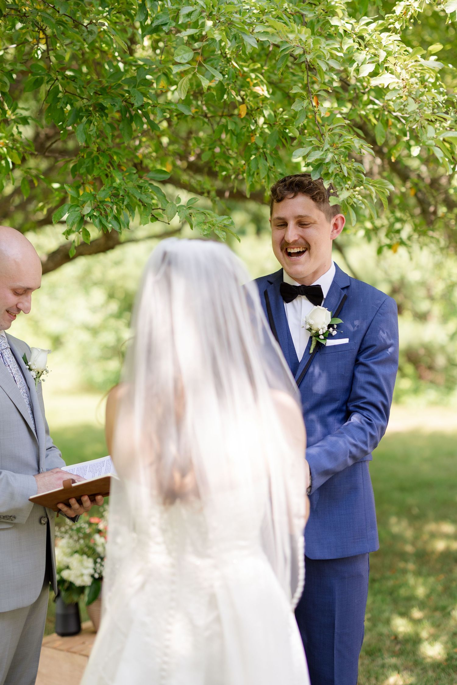 Groom in blue suit laughs joyfully during outdoor wedding ceremony under leafy green tree.
