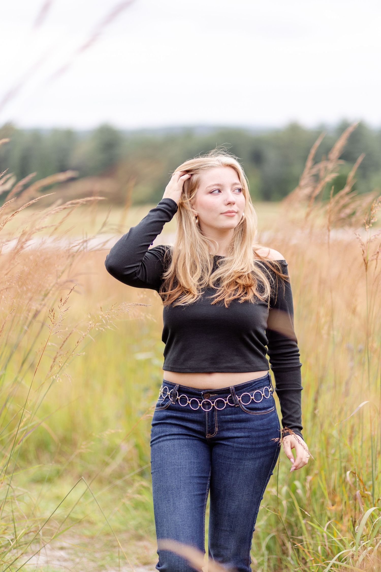 Blonde woman in black crop top and jeans walking through tall golden grass in open field.