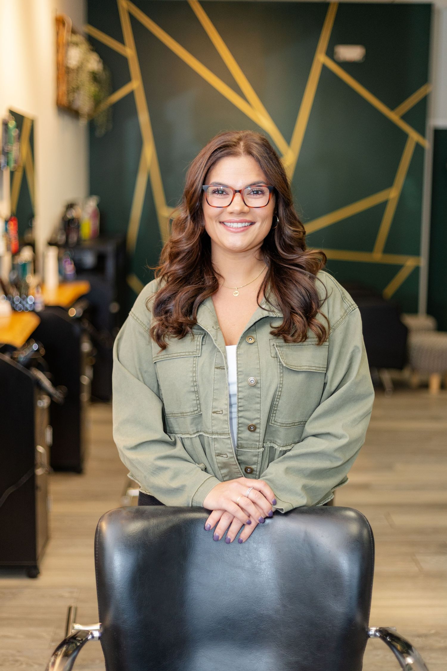 Smiling brunette hairstylist in olive jacket with glasses posing in green and gold salon.