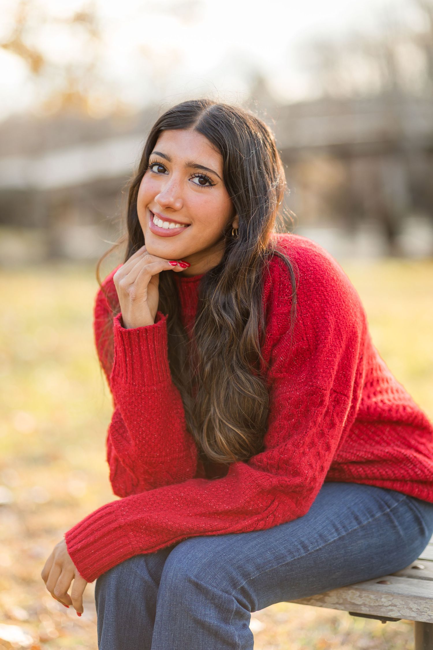 Young woman in red knit sweater and jeans sitting on bench smiling outdoors in fall.